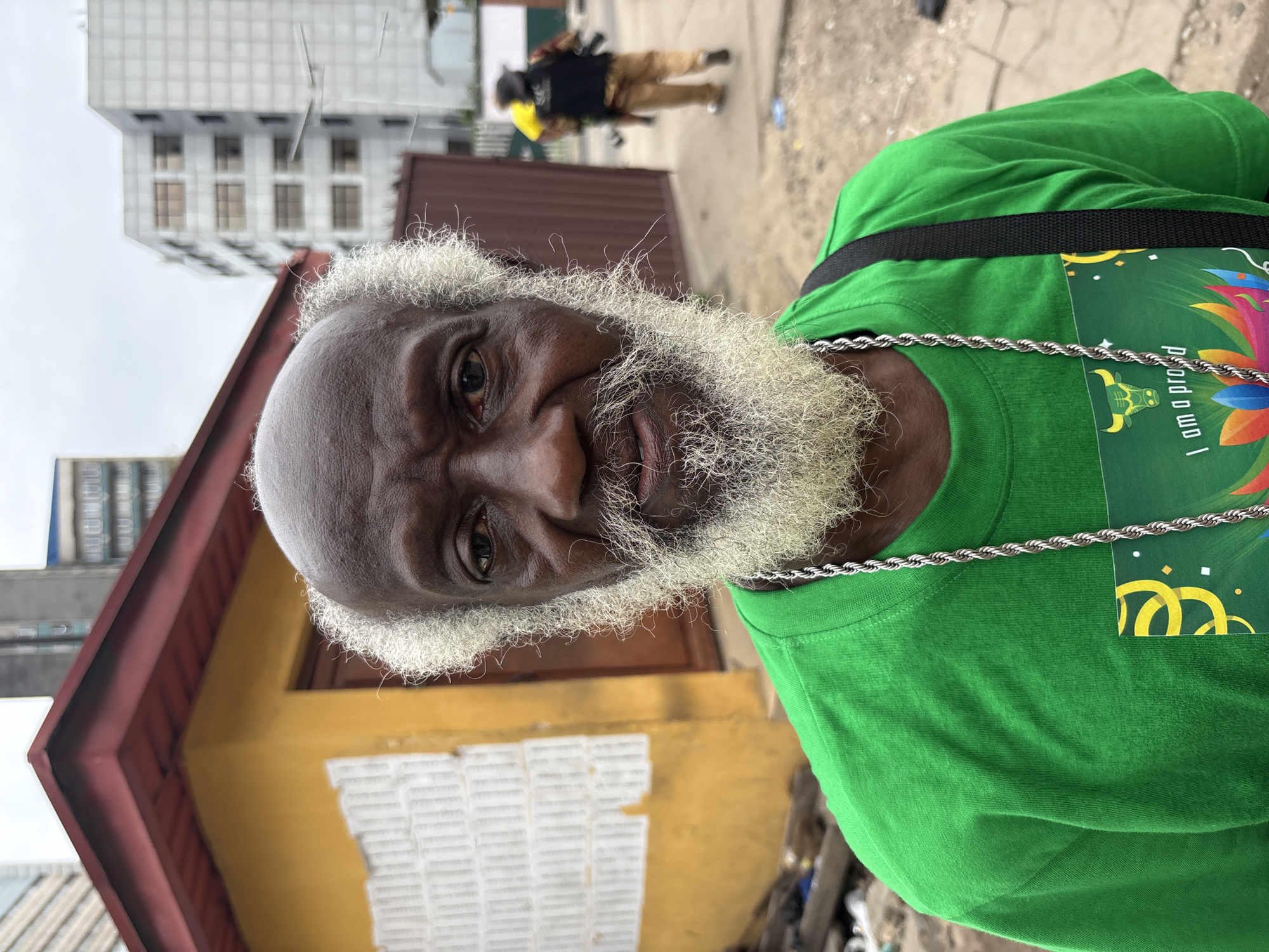 The image shows an elderly man with a dark complexion, balding head, and white hair and beard. He is wearing a green t-shirt with a graphic on the chest and a silver chain around his neck. A black strap crosses his shoulder. Behind him, there's a yellow building with a red roof and what appears to be papers pasted on the wall. In the background, there is a brown fence, a person walking away, a street, and a taller building. The image was taken in Lagos, Nigeria.