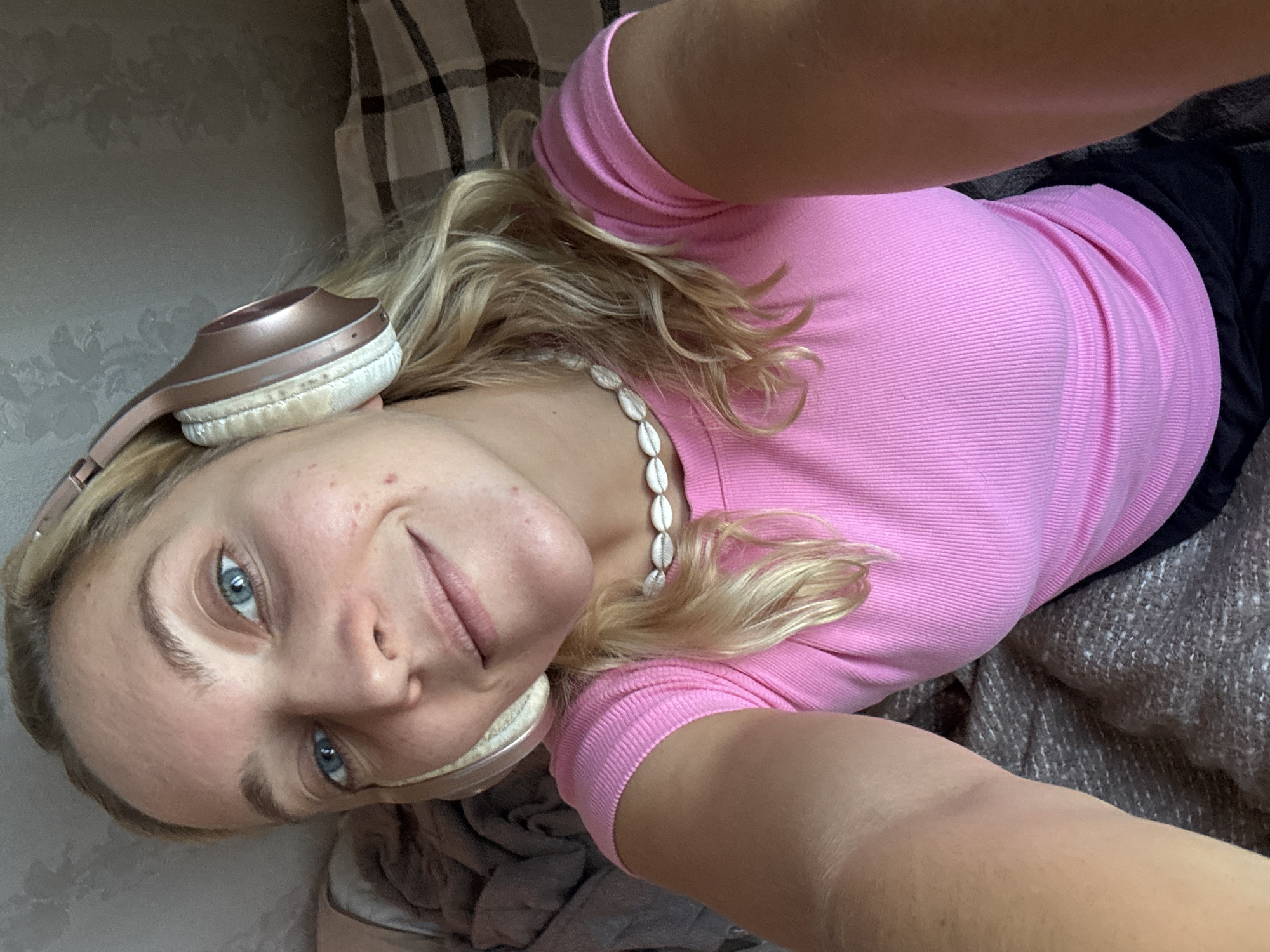 A young woman is captured in a close-up, upward-angled selfie, seemingly taken while she is lying down or reclining indoors. She has fair skin, blonde wavy hair, and blue eyes, which are directed towards the camera with a gentle, relaxed smile. A rose gold or bronze-colored pair of over-ear headphones rests partially on her head, with visible light-colored, possibly worn, earcups. Around her neck, she wears a necklace made of small white shells.
She is dressed in a short-sleeved, ribbed pink top, with a dark, possibly black, fabric visible on her lower body. The background consists of various textures: a light-colored plaid fabric in the upper left, a wall with a subtle patterned wallpaper (possibly floral or damask) behind her head, and a darker, textured fabric (like a blanket or throw) on the right side.
The lighting appears natural and bright, suggesting daytime. Her expression conveys a sense of calm and contentment. Minor skin imperfections are visible on her cheek and forehead. There is no visible text in the image. The scene suggests a casual and private moment of relaxation, possibly listening to music.