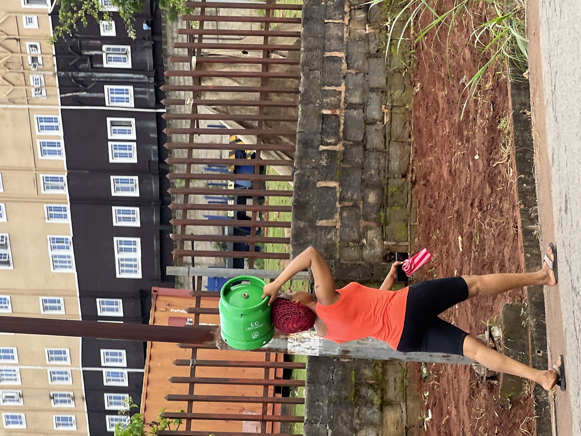 A woman walks along a paved path carrying a green propane cylinder on her head. She is wearing an orange tank top and black shorts, and she has a red head covering. A pink and white striped cloth is in her right hand. She is walking in front of a tall brick wall, which is behind a metal fence. Behind the fence are some buildings, including a tan and black building with many windows. The ground is covered in reddish-brown dirt and some sparse green grass. The image appears to be taken in Nkpor, Nigeria.