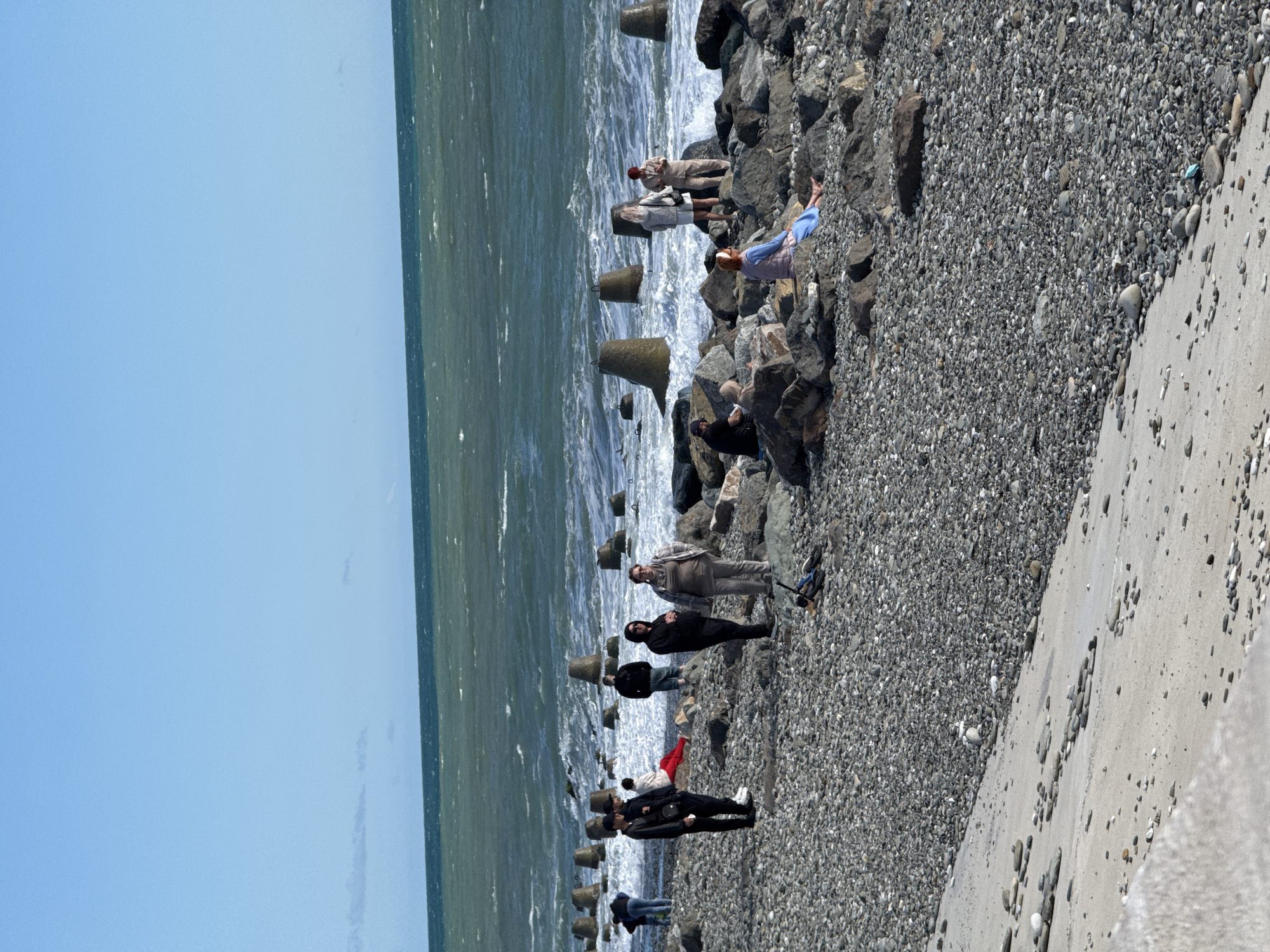 The image depicts a group of people standing on a rocky beach in Sirius, Russia. The beach is composed of small, gray stones and is bordered by a concrete wall. The sea is visible in the background, with waves crashing against a series of concrete tetrapods that act as wave breakers. A clear blue sky is visible.
Several individuals are scattered along the beach. Some are standing, while others are sitting or leaning against the rocks. Their clothing varies, with some wearing dark jackets and pants, while others are dressed in lighter colors. Some individuals are looking at the sea, while others appear to be conversing with each other. No specific actions or interactions beyond standing and conversing are evident in the image.