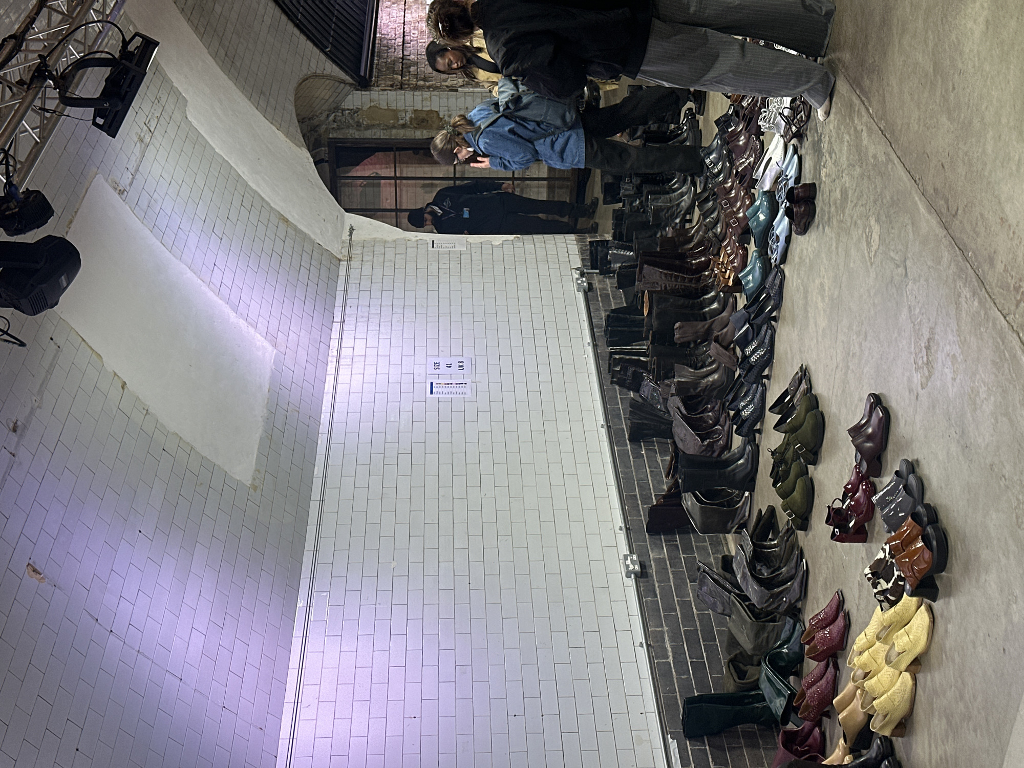 An indoor scene depicts an art installation or display of numerous pairs of shoes arranged on the floor. The floor is concrete, and the walls are covered with white rectangular tiles. Several people are visible in the background, observing the exhibit. The space appears to be in a large, enclosed area with a high ceiling. Lighting fixtures are visible. The location is in the city of London, United Kingdom.