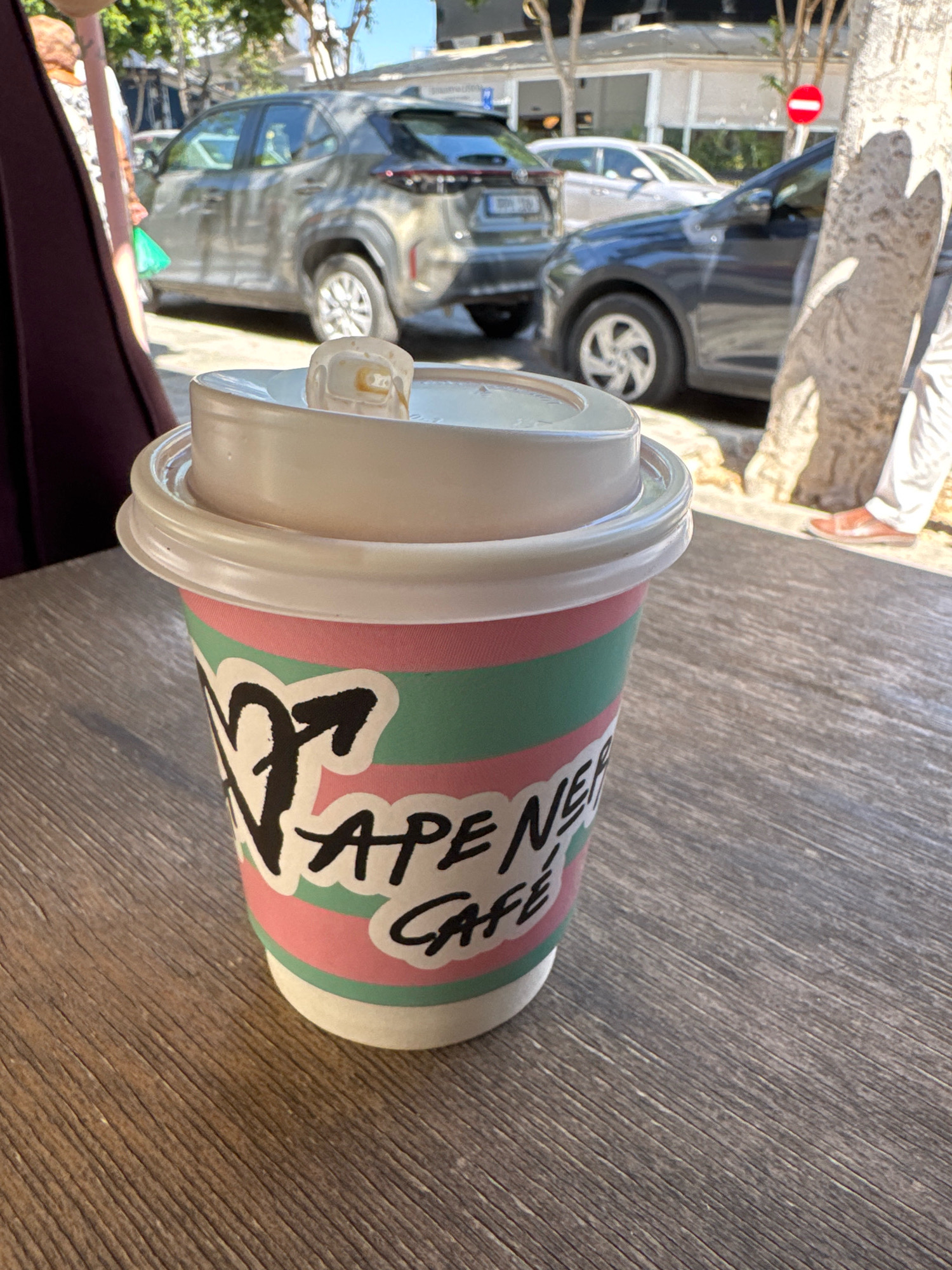 A close-up shot shows a disposable coffee cup with pink and green stripes and a white lid. The cup has black lettering that reads "VAPE NERV CAFE". The cup is placed on a wooden table outdoors. In the background, cars are parked on a street with trees and buildings. The scene appears to be in Limassol, Cyprus, given the presence of cars typical of the region and the general Mediterranean ambiance.