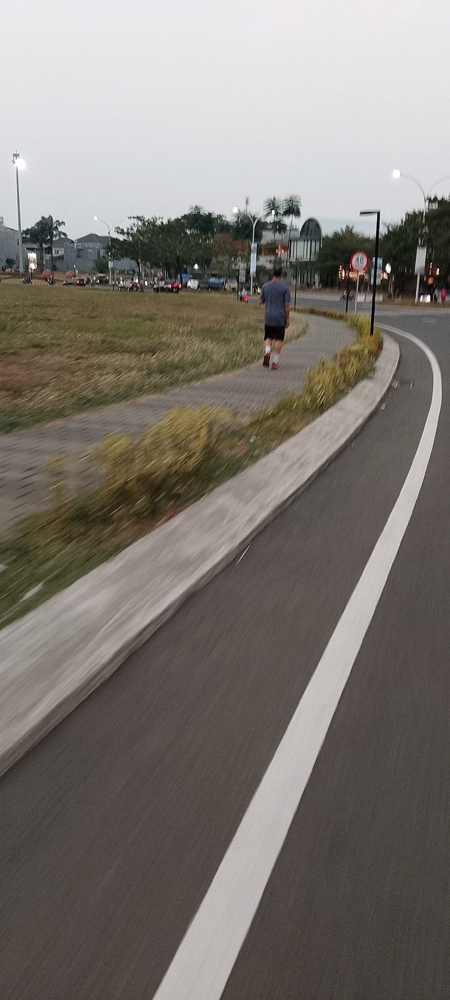 A person is walking on a paved path next to a grassy area. The person is wearing a blue shirt and black shorts with white socks and red shoes. They are walking away from the camera. The path curves to the right and is adjacent to a road with a white line marking. In the background, there are trees, buildings, and streetlights. A speed limit sign is visible in the background. The sky is overcast. The scene appears to be in a public park or recreational area in Gunung Putri, Indonesia.