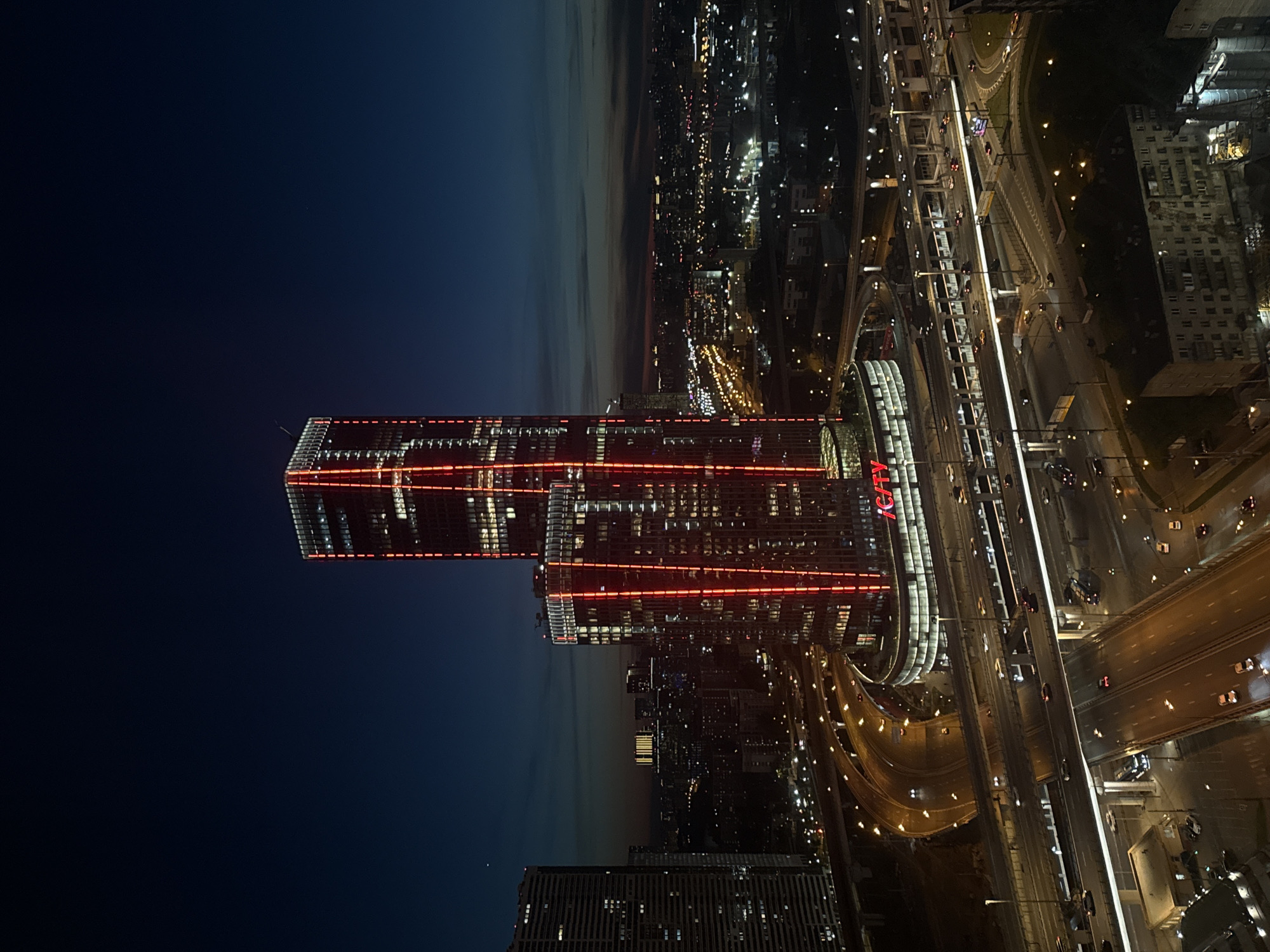 The image captures a vibrant urban landscape at night, viewed from a high vantage point. Two modern skyscrapers, prominently illuminated with bright red horizontal and vertical light strips, stand out on the left side of the frame. One building is notably taller than the other. Below these structures, a complex network of multi-lane highways, overpasses, and underpasses stretches across the midground and foreground, bustling with vehicular traffic. Cars are visible on the roads, their headlights and taillights creating streaks of light due to motion.
The scene depicts the ongoing activity of a city after dusk, with widespread artificial lighting illuminating the entire area. The sky in the background transitions from a deep twilight blue near the horizon to a dark, inky black overhead, indicating late evening or night. The multitude of lights from buildings, streetlamps, and vehicles creates a dynamic and expansive urban panorama.
Notable details include the striking red architectural lighting on the main buildings, which provides a distinctive visual signature. The high-angle perspective offers a comprehensive view of the city's infrastructure and expanse.
Visible text includes the red illuminated word "CITY" on the base of the larger, rightmost prominent building. To its left, on the base of an adjacent building, the Cyrillic word "ХИМКИ" (Khimki) is also visible in red. On the base of the tallest skyscraper on the far left, partially obscured, the Cyrillic text "АРК" (ARK) can be discerned. The location context indicates this scene is in Khimki, Russia.