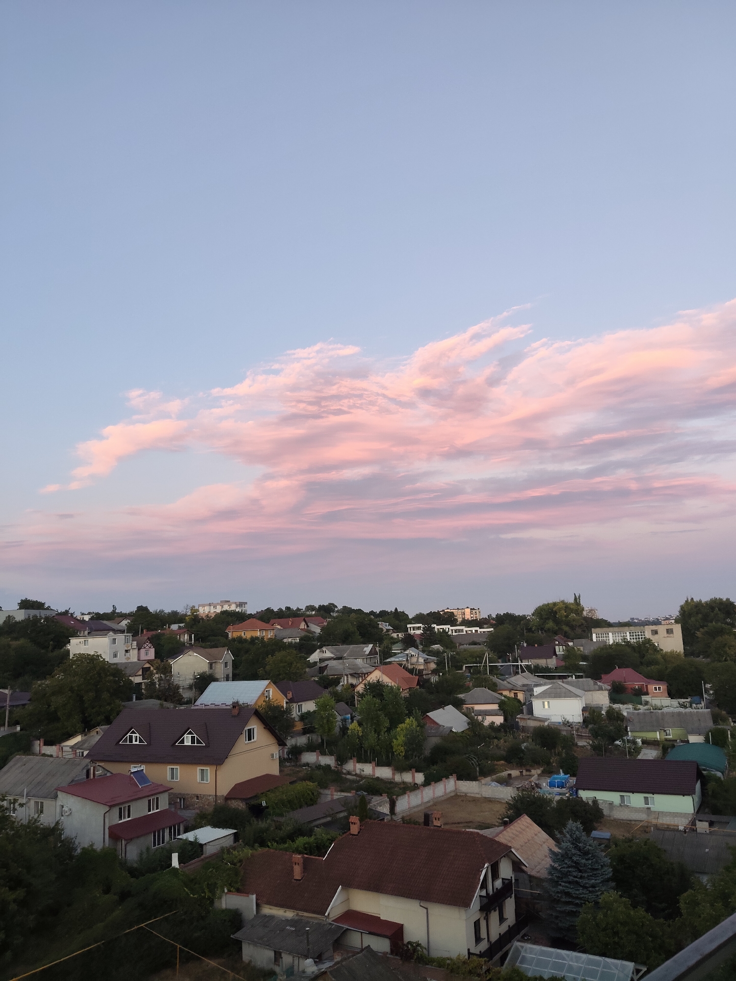 This is a high-angle, wide shot of a residential area in Durlești, Moldova, at dusk. The sky is a pale blue with streaks of pink and orange clouds, suggesting the setting sun. Below, a densely packed neighborhood of houses with various roofing materials and architectural styles is visible, interspersed with lush green trees. The houses are mostly two-story structures, with some single-story buildings and garages. Fences and walls delineate property lines. There are no people or visible activity captured in the image. The overall atmosphere is calm and suburban.
