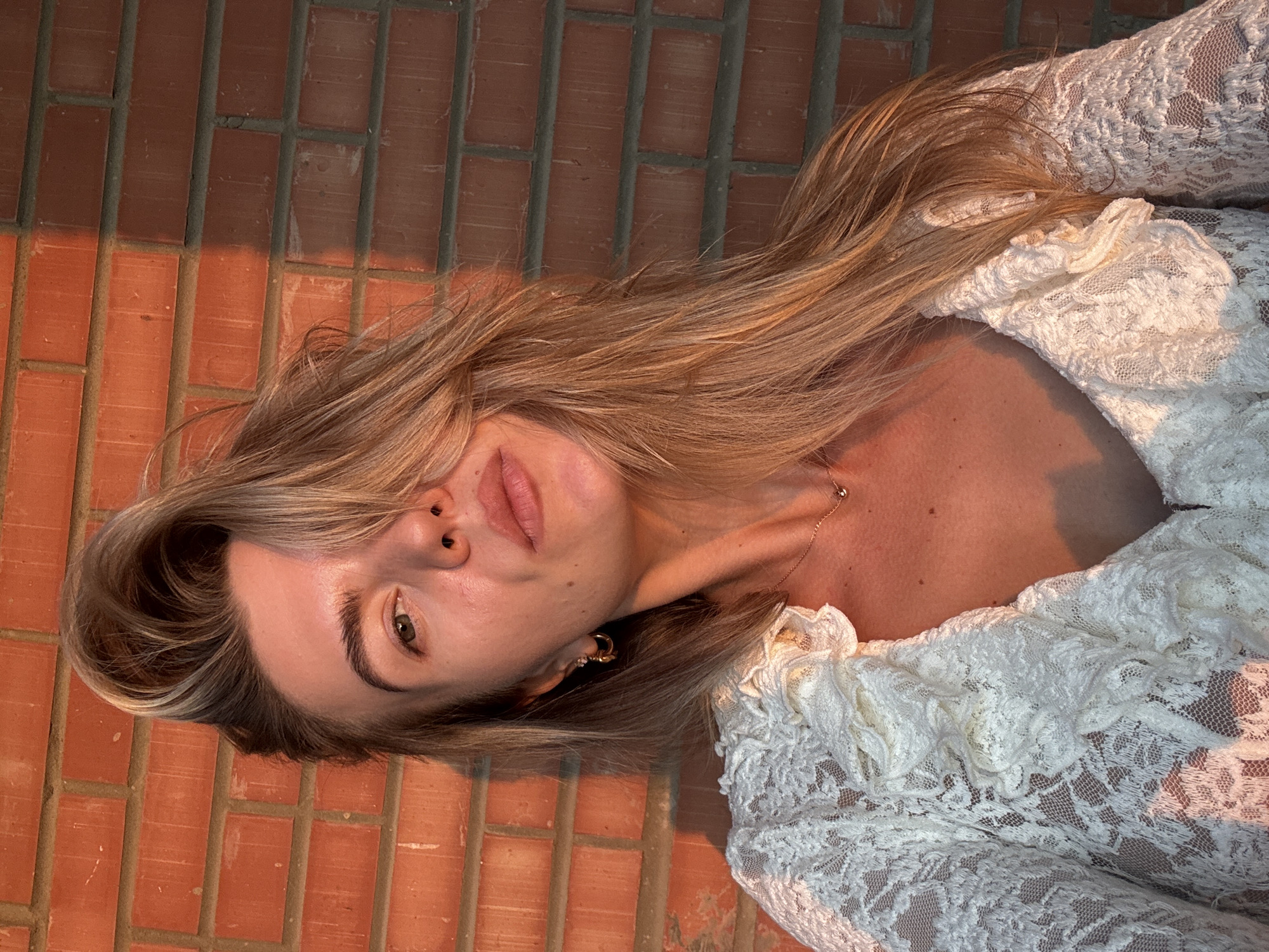 A young woman with blonde hair is looking at the camera. She is wearing a white lace top with ruffles. The background is a brick wall, with a shadow cast on the bricks. The lighting suggests it is either sunrise or sunset.
