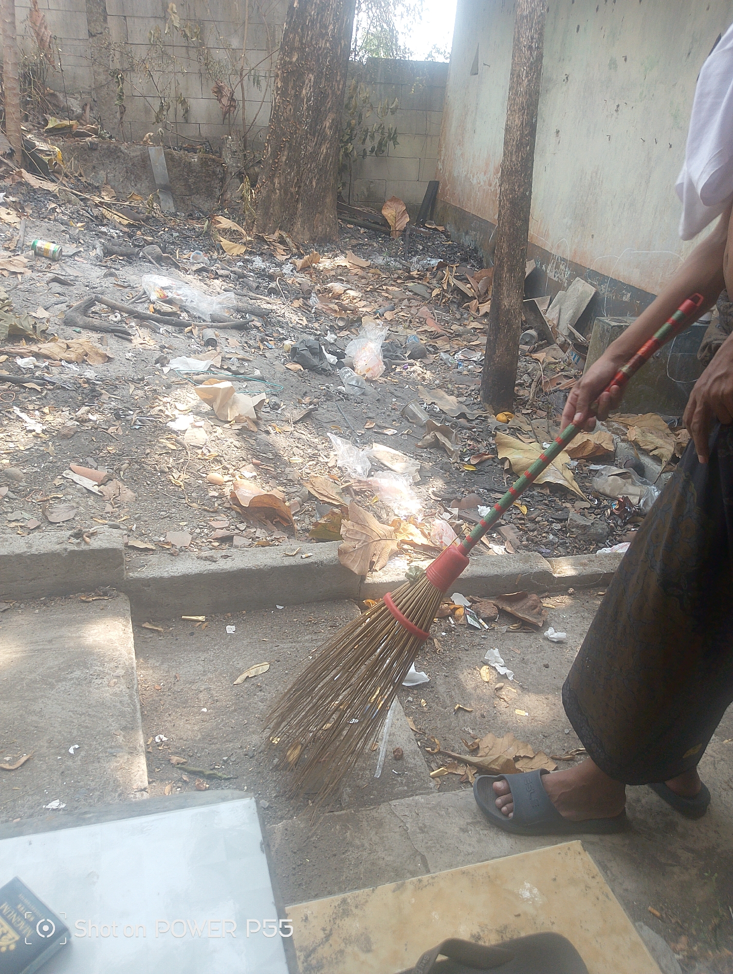 A person is sweeping a littered outdoor area in Lebaksiu, Indonesia. The ground is covered with dry leaves, dirt, and various types of trash, including plastic bags, bottles, and discarded wrappers. A broom with a red and green striped handle is being used to gather the debris. The person is wearing a dark patterned sarong and dark sandals. In the background, a rough concrete wall and a tree trunk are visible. The overall scene depicts a neglected and unkempt environment.