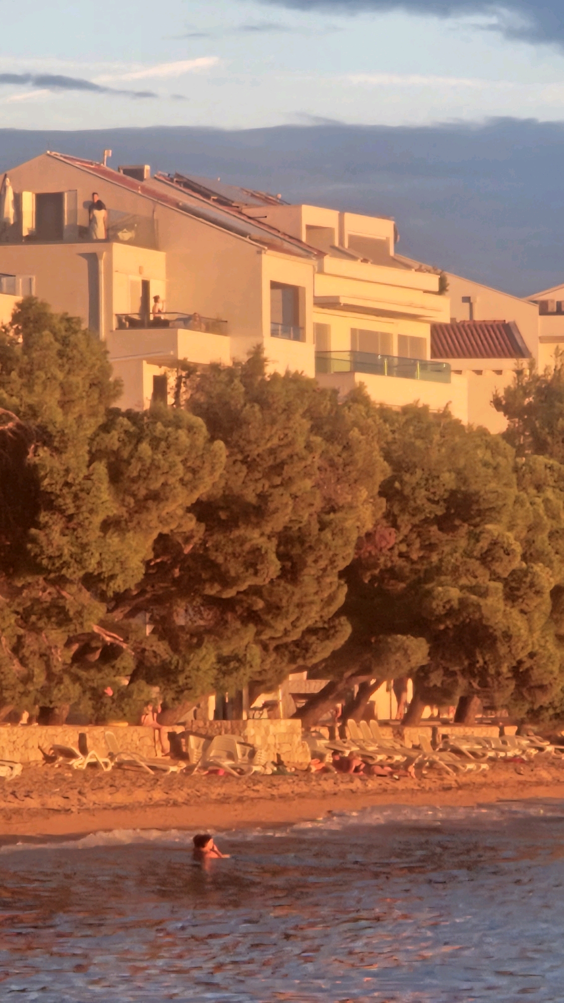 The image depicts a coastal scene at sunset, likely in Općina Tučepi, Croatia. In the foreground, the sea reflects the warm glow of the setting sun, with gentle waves lapping at the shore. A person is visible in the water, partially submerged. The beach area is lined with numerous empty lounge chairs, suggesting a quiet moment before or after peak activity. Behind the beach, a dense line of green trees forms a natural barrier, with their branches reaching towards the sky. Above the trees, a modern, multi-story building complex is visible, featuring balconies and rooftop elements, some of which appear to have solar panels. Two individuals are standing on a balcony of this building, and another person is visible on a lower balcony. The sky above displays a gradient of colors, transitioning from a soft blue to darker clouds, indicative of twilight. The overall impression is one of a peaceful, sun-drenched evening at a resort or residential area by the sea.