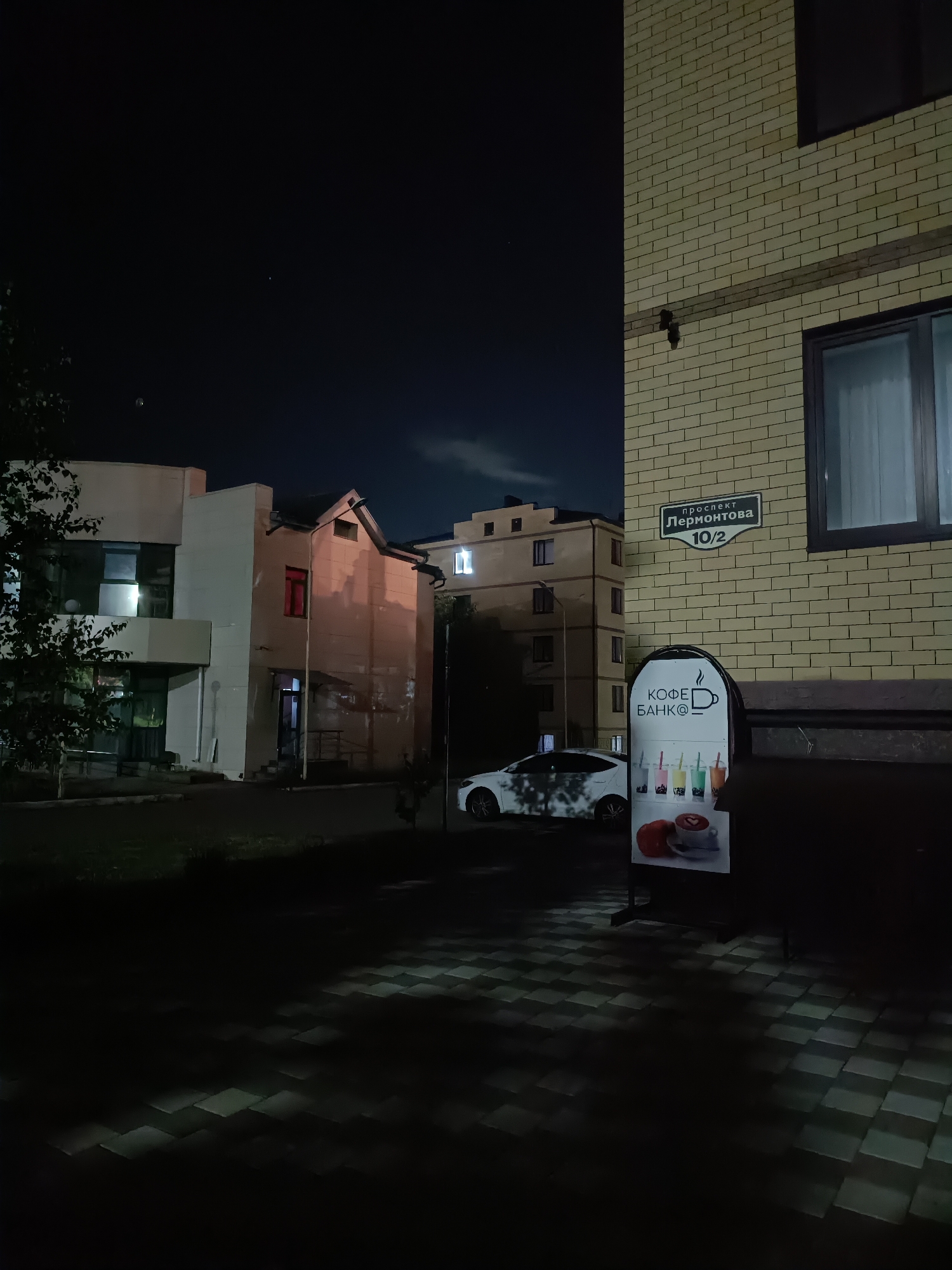 The image depicts a night scene at 10/2 Lermontov Avenue in the city of Lermontov, Russia. The overall environment is an urban outdoor area, illuminated by artificial light sources under a dark night sky. A faint, diffuse cloud formation is visible in the upper right portion of the sky, with a single faint light point in the upper left.
On the right side of the frame, a multi-story building constructed of light-colored, possibly pale yellow or beige, brick is prominent. A dark horizontal band of bricks extends across its facade. A dark-framed window is visible, with a light-colored curtain discernible within. Attached to this building is a dark rectangular street sign with white Cyrillic text, reading "проспект Лермонтова" (Lermontov Avenue) and "10/2" below it.
In the right foreground, in front of the brick building, a standalone A-frame commercial sign is positioned on the paved ground. The sign features a coffee cup icon and the Cyrillic text "КОФЕ БАНК@" (Coffee Bank@) at the top. Below this, there are illustrations of several colorful beverages in clear cups, and at the bottom, an illustration of a coffee cup with heart-shaped latte art alongside a pastry.
To the left of the frame, a lighter-colored building, possibly white or pale grey, with a more modern architectural style and large reflective windows, is partially visible. Dark tree foliage partially obscures the lower left portion of this building. Further in the background, another multi-story building with illuminated windows is discernible. A white sedan is parked on the dark asphalt road in the midground, its surface reflecting ambient lights.
The immediate foreground features a paved surface composed of light and dark square pavers arranged in a checkerboard pattern, reflecting scattered light. Dark ground cover, possibly grass, is present in the lower left. No individuals are visible in the scene.