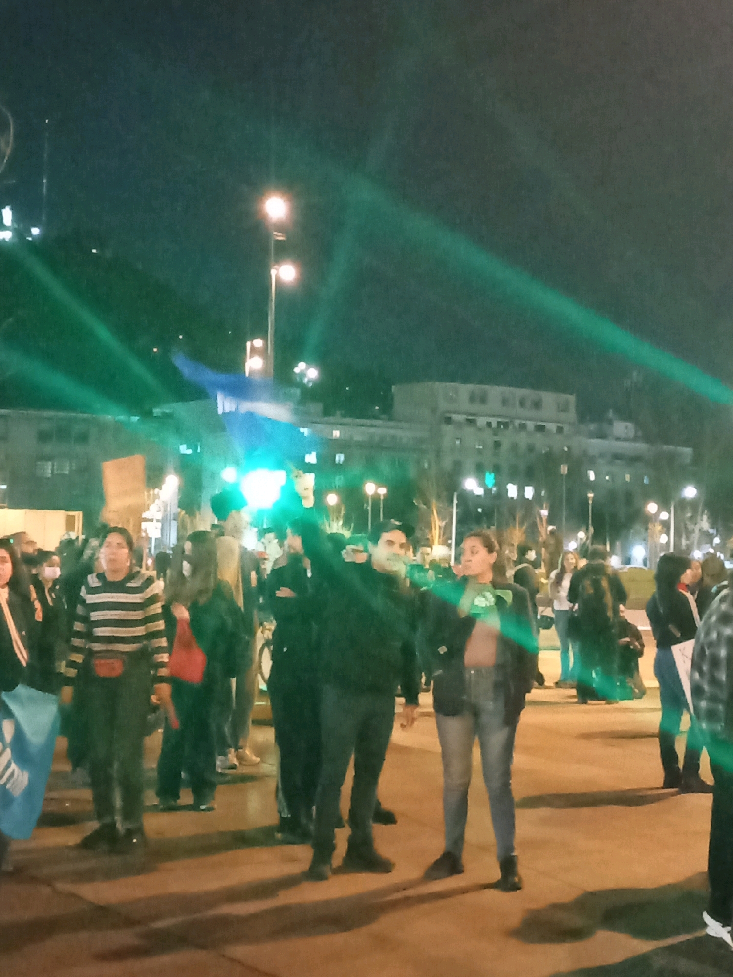 The image depicts an outdoor nighttime scene in the city of Providencia, Chile. A group of people are gathered in what appears to be a public square or plaza, with a large, multi-story building visible in the background, illuminated by lights. Several streetlights are also present, casting light on the scene and creating lens flares that streak across the image. The ground appears to be paved.
In the foreground, several individuals are standing and walking. One person on the left is wearing a black jacket and black pants and is holding a blue and white flag. To their right, a person in a horizontally striped sweater and dark pants is visible. Behind them, a person in a dark jacket and dark pants is holding a protest sign. Further back, a group of people are standing and interacting. One individual in the center is wearing a green shirt and blue jeans. To their right, another person in a dark jacket and blue jeans is also visible. Some people are wearing face masks.
The overall atmosphere suggests a public gathering or demonstration, possibly a protest, given the presence of signs and flags, and the nighttime setting. The green streaks across the image are likely from lighting effects or camera artifacts.