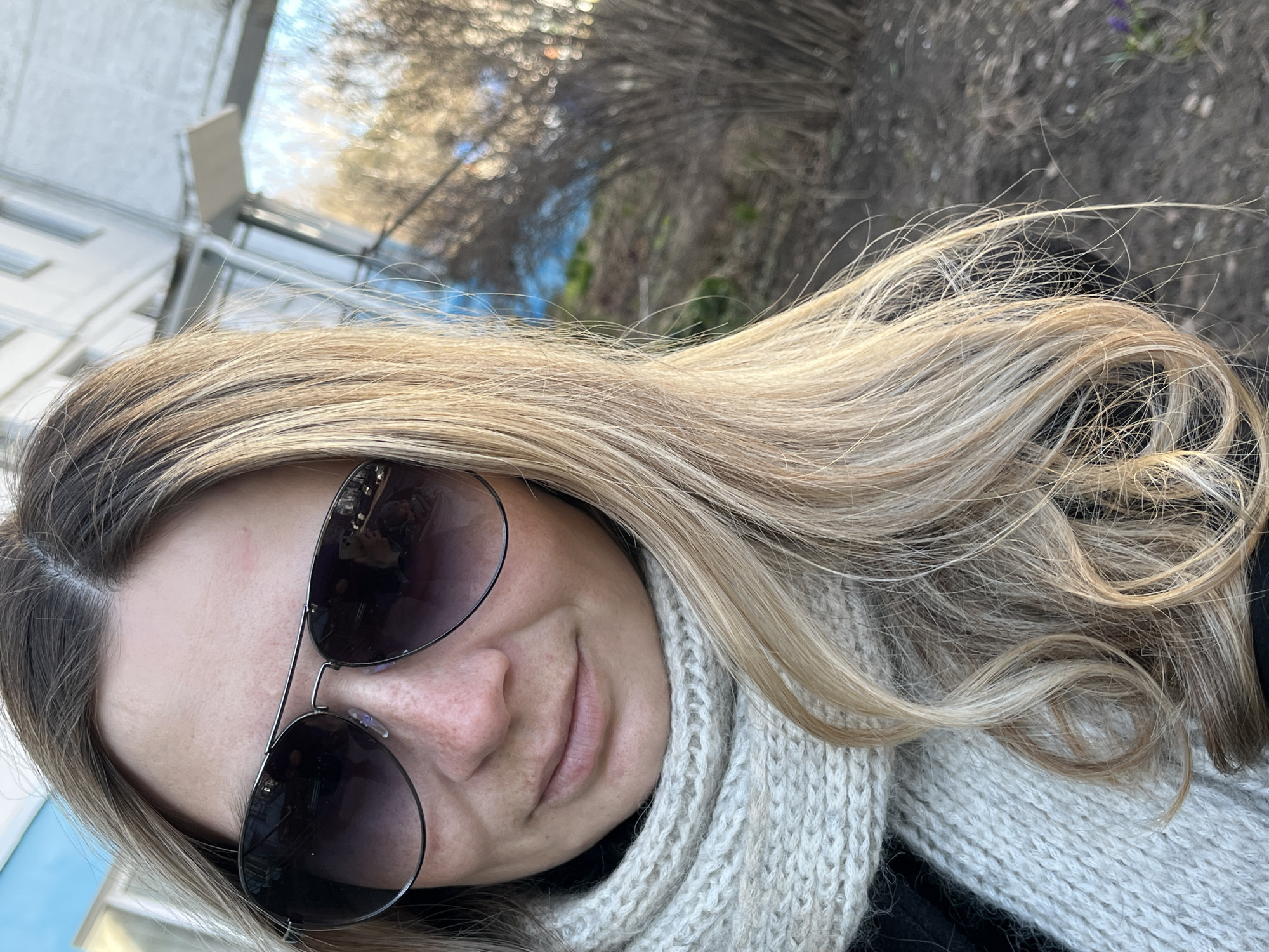 The image is a close-up portrait of a woman with long, blonde hair. She is wearing sunglasses and a knitted scarf. The background is blurry and appears to be outdoors, with trees and buildings visible. The location is Chaikovsky, Russia.