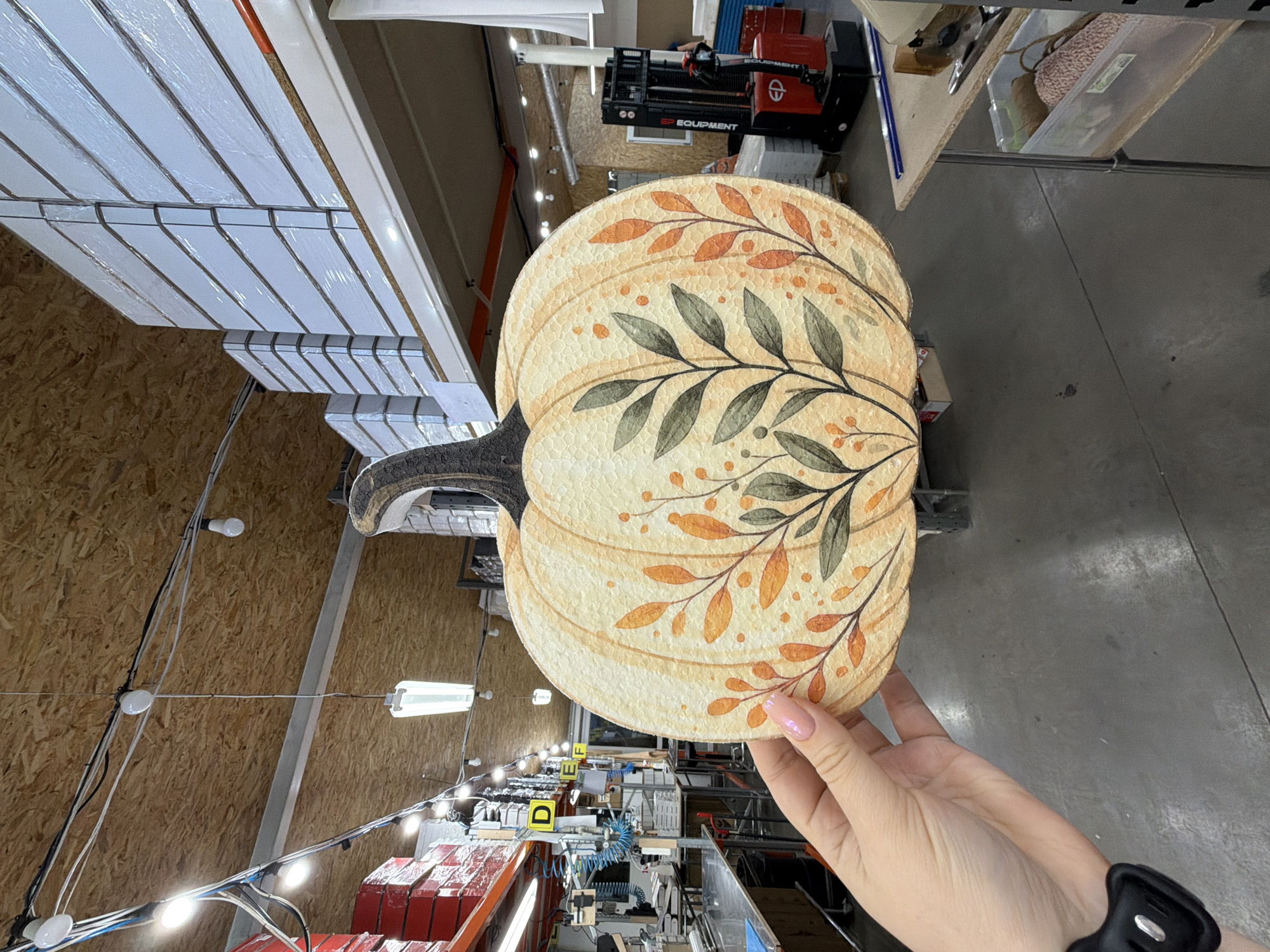 A close-up, low-angle medium shot shows a woman's hand with light pink nail polish holding up a cream-colored, flat pumpkin decoration adorned with painted autumn leaves and branches against a background of an indoor warehouse. The pumpkin has a textured surface and a dark brown stem. The warehouse environment features a concrete floor, industrial shelving stacked with white boxed items wrapped in plastic, red boxes, fluorescent lighting fixtures suspended from a wood-paneled ceiling with exposed wires, and a red and black forklift in the mid-ground. The overall lighting is neutral, highlighting the pumpkin decoration.