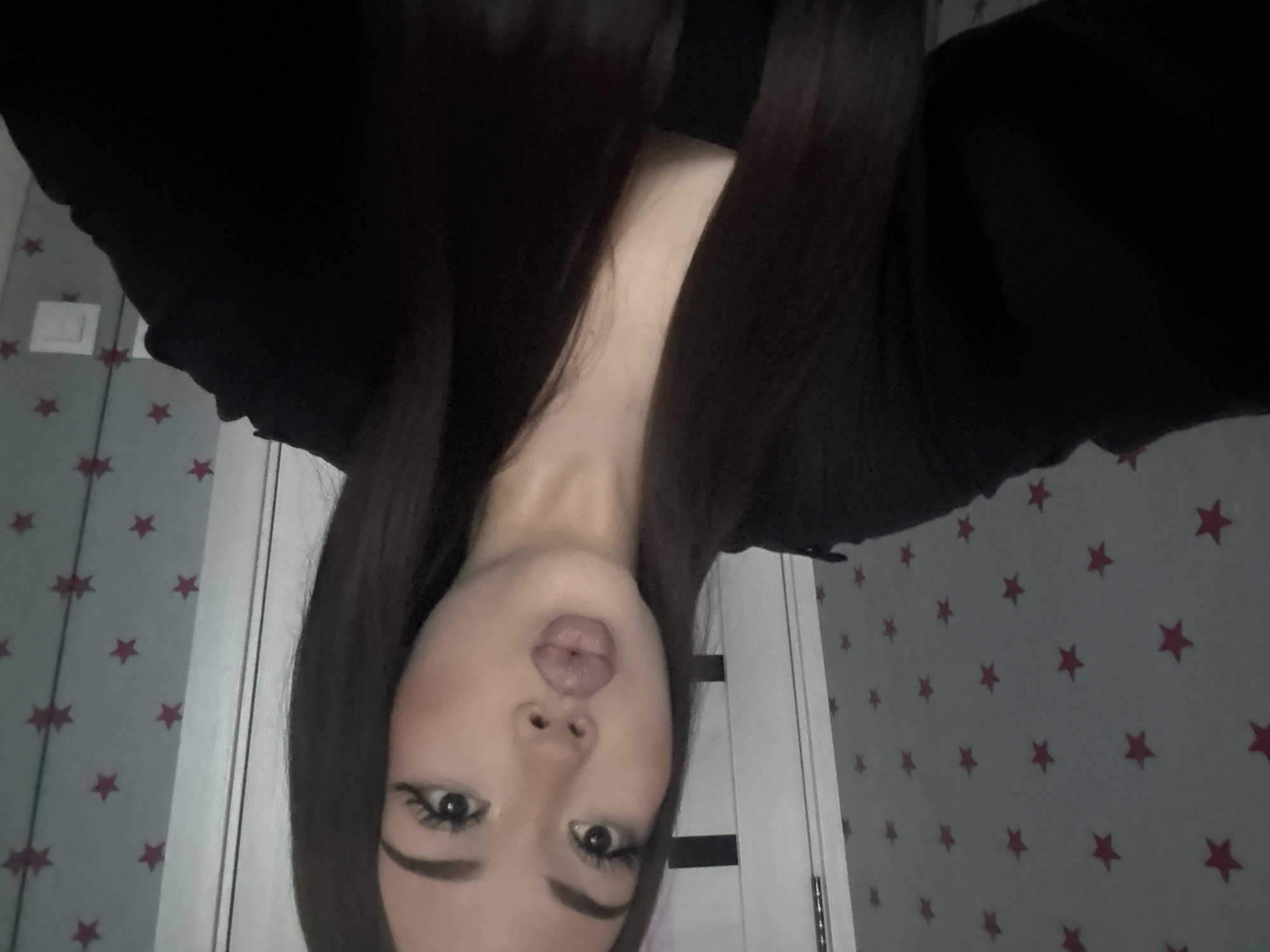 A young woman with long dark hair and heavy makeup is taking a selfie in a room with a star-patterned wallpaper. She is wearing a black top. The background includes a door and a reflection in a mirror showing more of the room. The image appears to be taken indoors. There is no information available that places this image in the city of Naberezhnye Chelny, Russia.