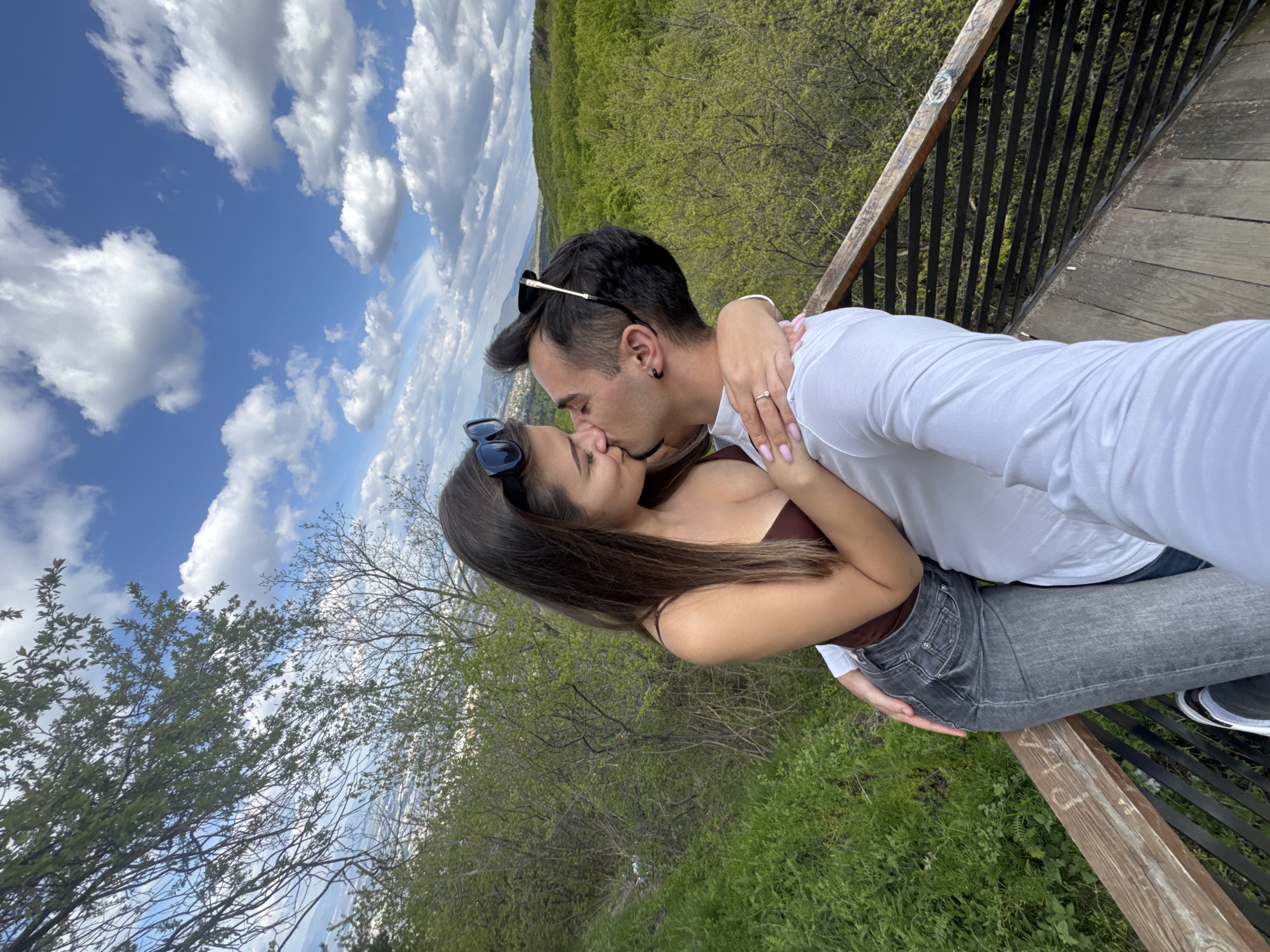 A couple is kissing on a wooden platform overlooking a landscape. The woman has long brown hair and is wearing a brown top and gray jeans. Sunglasses are perched on her head. She is embracing the man. The man has short dark hair and is wearing a white shirt and jeans. He has sunglasses on his head. The sky is blue with white clouds. Green trees cover the landscape. The setting could be near Sofia, Bulgaria, given the vegetation and potential distant cityscape.