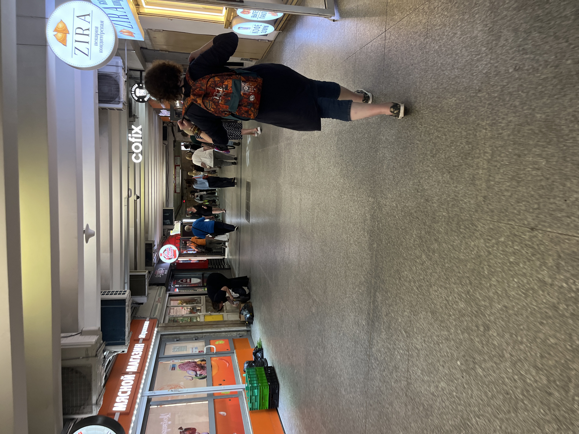 The image depicts a pedestrian underpass or walkway in Minsk, Belarus, likely a commercial area. In the foreground, a woman with dark, curly hair, wearing a dark dress and blue jeans, walks away from the camera, carrying a colorful backpack. Her shoes are black and white patterned slip-ons. To her left, further down the walkway, a person in a black outfit is bent over, rummaging through several black trash bags and green crates.
The walkway has a light gray, speckled floor and a white, paneled ceiling with visible air conditioning units. Numerous shops and businesses line the sides of the walkway, indicated by signs. To the left, a shop with the Cyrillic text "МЯСНОЙ МАГАЗИН" (Meat Shop) is visible. Further down, a sign for "cofix" is illuminated. On the right, a circular sign with Cyrillic text and an orange symbol reads "ZIRA пекарня кондитерская" (ZIRA bakery confectionery). Other businesses are also present, with visible signage in Cyrillic. People are seen walking in the background, suggesting a public thoroughfare. The lighting appears to be artificial, consistent with an indoor or underground setting.