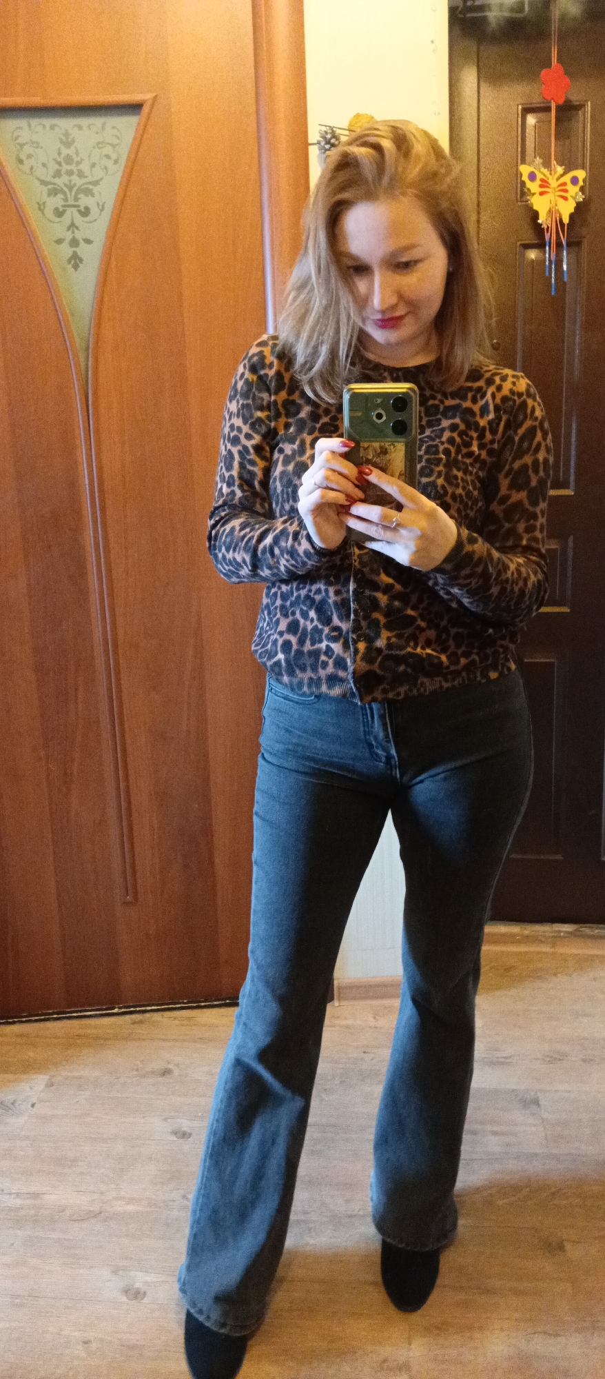 A woman is captured in a mirror selfie, standing indoors in what appears to be a hallway or room. She has light brown hair, parted on the side and falling loosely around her shoulders, and wears red lipstick. Her attire consists of a brown and black leopard print button-up cardigan and dark grey or blue flared jeans. Her fingernails are painted red, and she wears a ring on her left ring finger. She holds a dark-cased smartphone with multiple camera lenses in both hands, directing her gaze downwards towards the screen.
The background features two wooden doors and a light wood laminate floor. To her left, there's a lighter wooden door with a distinctive decorative patterned glass panel. To her right, a darker wooden door is adorned with a hanging decoration that includes a red flower and a yellow butterfly with blue accents. A light-colored wall is visible above the doors. The overall lighting suggests an indoor, possibly daytime, setting with natural light. The woman displays a soft, subtle smile as she takes the photograph. No discernible text is visible in the image.