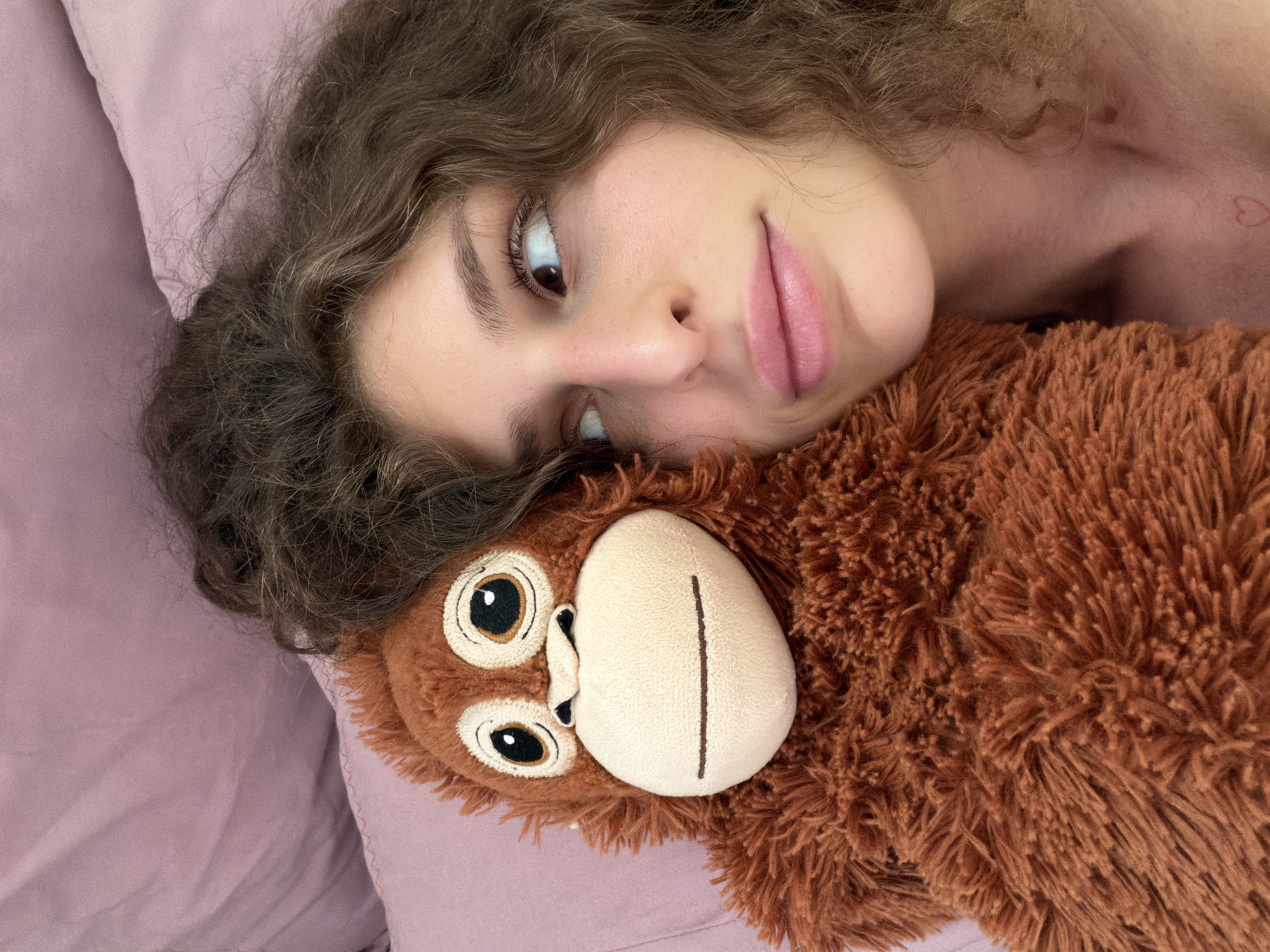 The image shows a person lying down with their head resting on a brown, stuffed monkey toy. They are on a lavender-colored pillow. The person has curly brown hair, and the skin on their face and visible shoulder is light-toned. A small, red heart tattoo is visible on the shoulder. The individual's gaze is directed slightly upwards and to the left. The monkey toy has large, dark eyes and a light-colored face.
There is no information to suggest the image was taken in North Kuta, Indonesia.