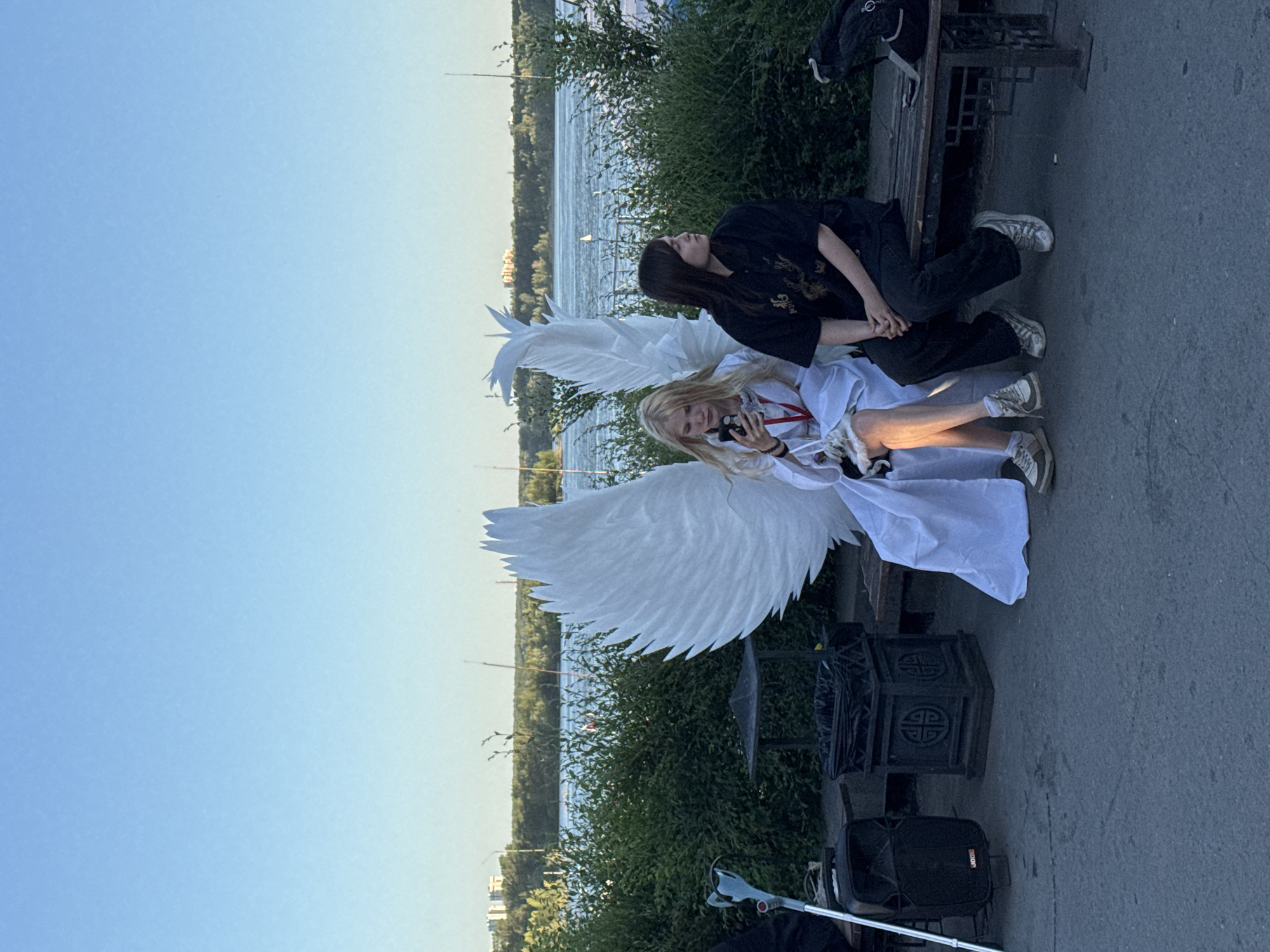 Two young women are seated on a park bench beside a body of water. The woman on the left is dressed in a white, flowing robe and has large white angel wings attached to her back. She is looking at her phone and holding a small white animal, possibly a cat. The woman on the right is wearing a black shirt and dark pants, and she is looking away from the camera. The background shows a wide river with a tree line and buildings on the opposite bank. The foreground is a paved area. A speaker and a cane are visible to the left of the bench. The scene appears to be set in a public park in Novosibirsk, Russia, given the presence of the river and typical park amenities.