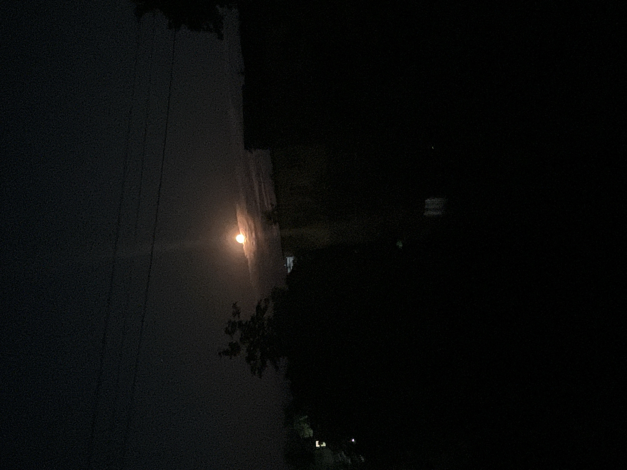 The image depicts a nighttime scene, likely in Poltava, Ukraine, dominated by darkness. A bright, full moon is prominently visible in the upper center of the frame, partially obscured by wispy clouds. A beam of light emanates from the moon, cutting through the night sky. Silhouettes of trees and buildings are visible against the dark backdrop, suggesting an urban or suburban environment. The lower portion of the image is almost entirely black, with faint hints of indistinct shapes and possibly distant lights. There are no discernible individuals or clear actions taking place in the scene. The overall atmosphere is one of quiet, nocturnal ambiance.
