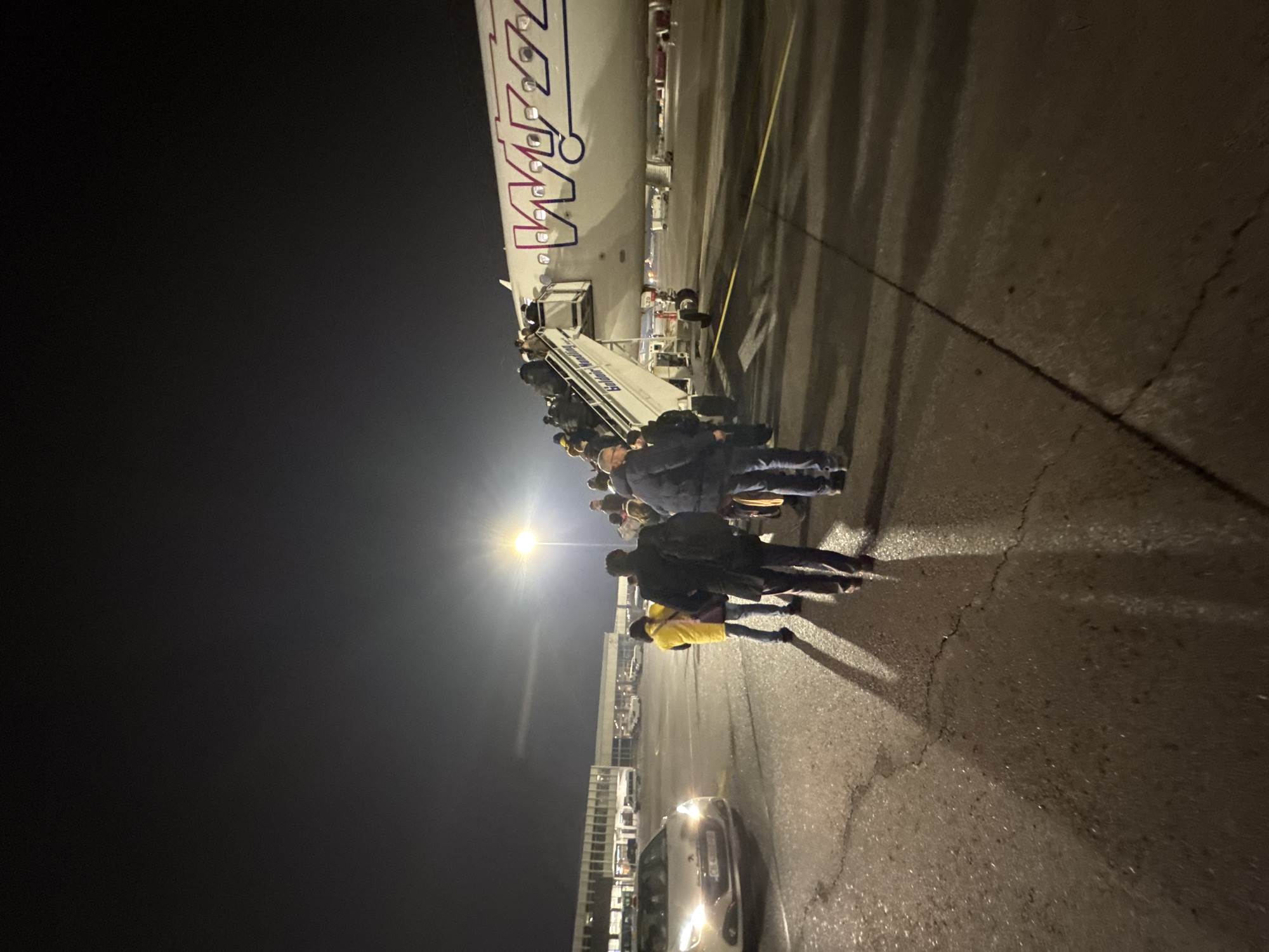 At night, passengers are disembarking a Wizz Air plane using a mobile stairway. The location is likely Sofia Airport in Sofia, Bulgaria, given the airline's presence there. A car is parked nearby on the tarmac. People are standing on the tarmac, their shadows cast by a bright light source. A building is visible in the background. The sky is dark. The scene is lit by artificial lighting.