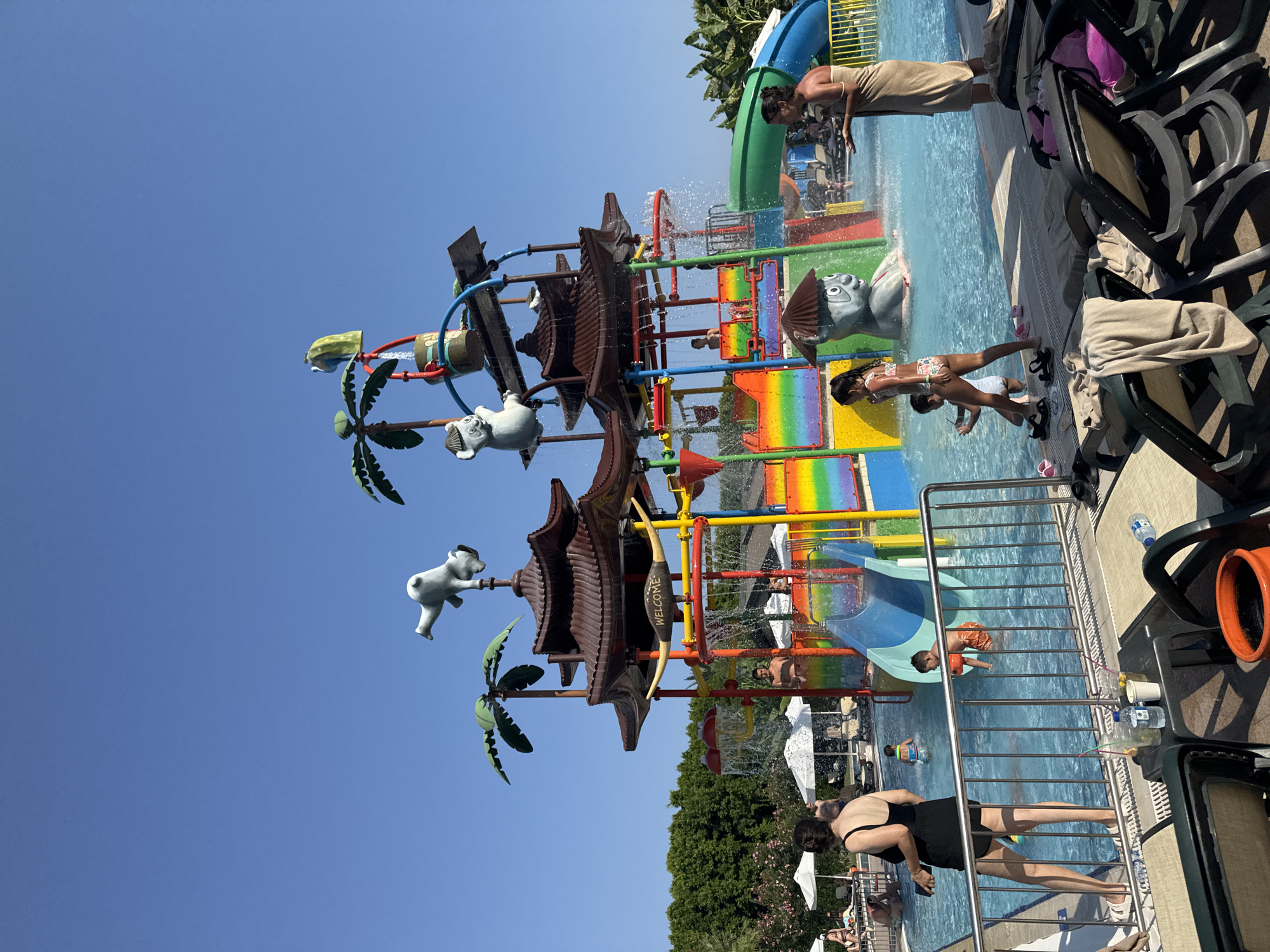 In Aksu, Turkey, a sunny day is captured at a water park. A large, multi-level water play structure dominates the scene, featuring colorful slides, water cannons, and decorative elements like palm trees and cartoon animal figures. Water cascades down from various points on the structure, creating a lively atmosphere. A swimming pool is visible in the foreground, with several children and adults enjoying the water.
In the foreground, a woman in a black swimsuit stands by the pool, holding a mobile phone. To her right, a young boy in orange swim trunks and a life vest is partially submerged in the water. Further into the pool, a girl in a patterned swimsuit is walking, and behind her, a young child is playing. To the right of the water structure, a woman wrapped in a towel stands in the water.
The surrounding area includes lounge chairs with towels draped over them, indicating a relaxing poolside environment. Lush green trees form a backdrop to the water park, and a clear blue sky suggests pleasant weather. Umbrellas are also visible, providing shade for guests. The overall impression is one of a family-friendly vacation destination.