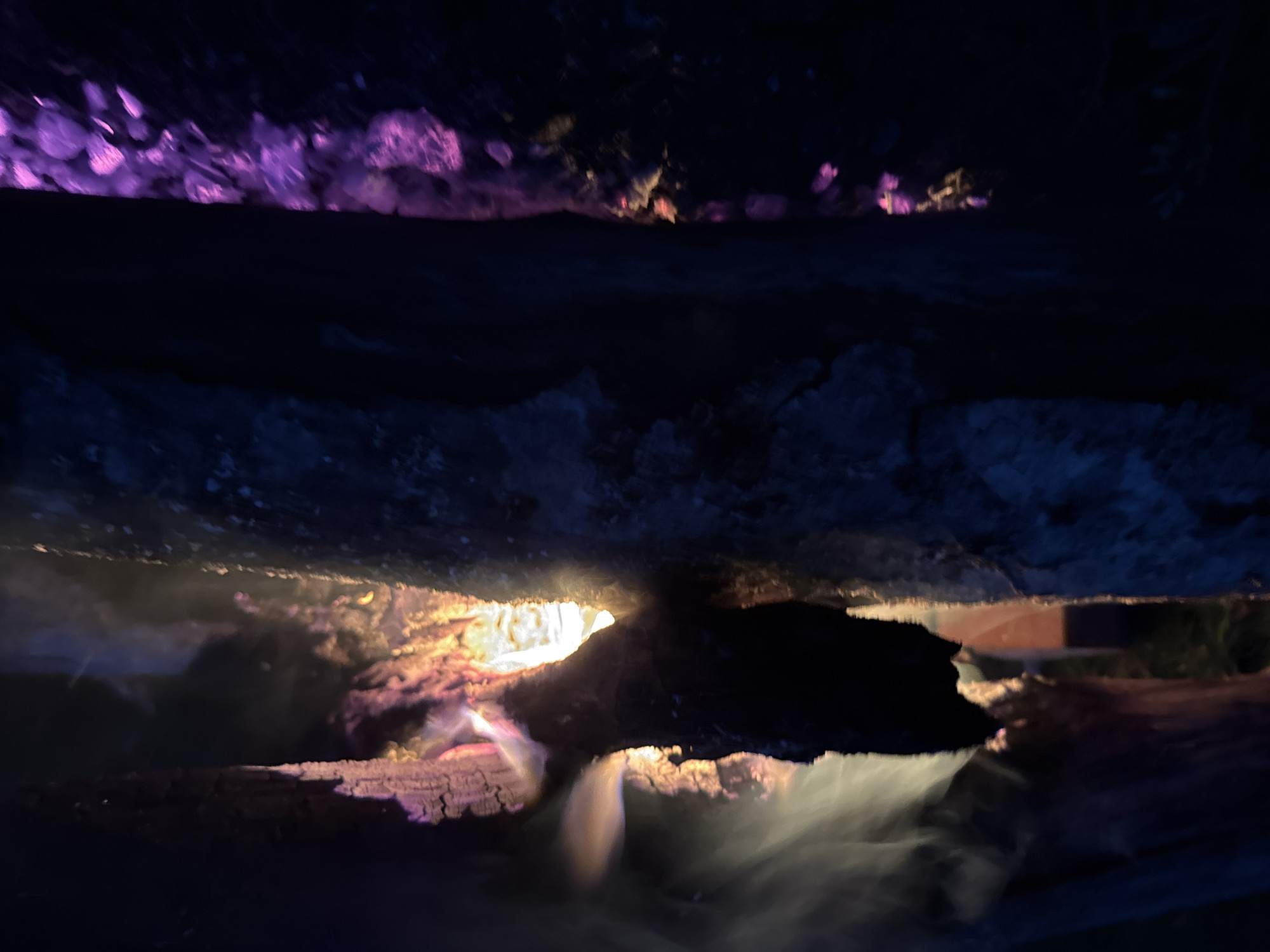 The image depicts a close-up, low-angle shot of burning logs at night. The logs are arranged horizontally, with smoke and flames rising from the top log, creating an ethereal glow. The fire's light illuminates the charred wood, revealing textures and cracks. Below the burning logs, there's a dark, indistinct surface that appears to reflect the fire's colors, with a notable purple hue in the lower portion of the frame. The overall scene is dark and atmospheric, suggesting an outdoor setting, possibly a campfire or fireplace. There is no visible indication of a specific location within the city of Ardu, Estonia. The image focuses solely on the natural phenomenon of fire and burning wood.