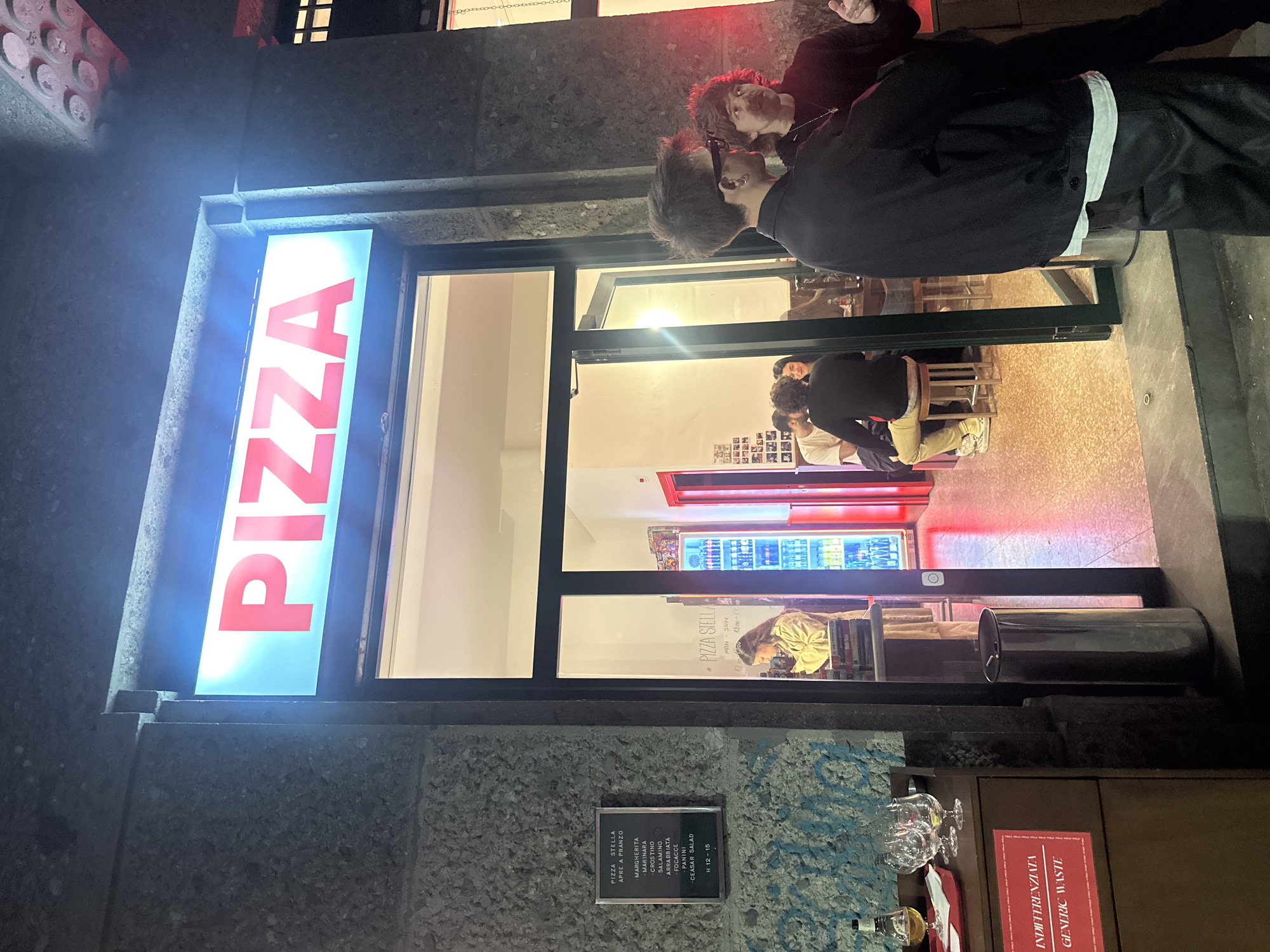 The image captures the exterior of "PIZZA STELLA", a pizzeria in Milan, Italy. The bright neon sign "PIZZA" is prominently displayed vertically. A man with dark hair and glasses is standing near the entrance, interacting with another individual whose face is partially visible.
Inside the pizzeria, there are other people seated at tables. A red-framed refrigerated display case filled with beverages is visible. A sign reading "PIZZA STELLA APRE A PRANZO" is on the building's exterior, listing pizza varieties. Another sign reads "*PIZZA STELLA MON - SUN 12.00 - 15.00 / 18.00-00.00". Below the menu, a sign indicates "INDIFFERENZIATA GENERIC WASTE". Several wine glasses and bottles are seen near the waste sign.