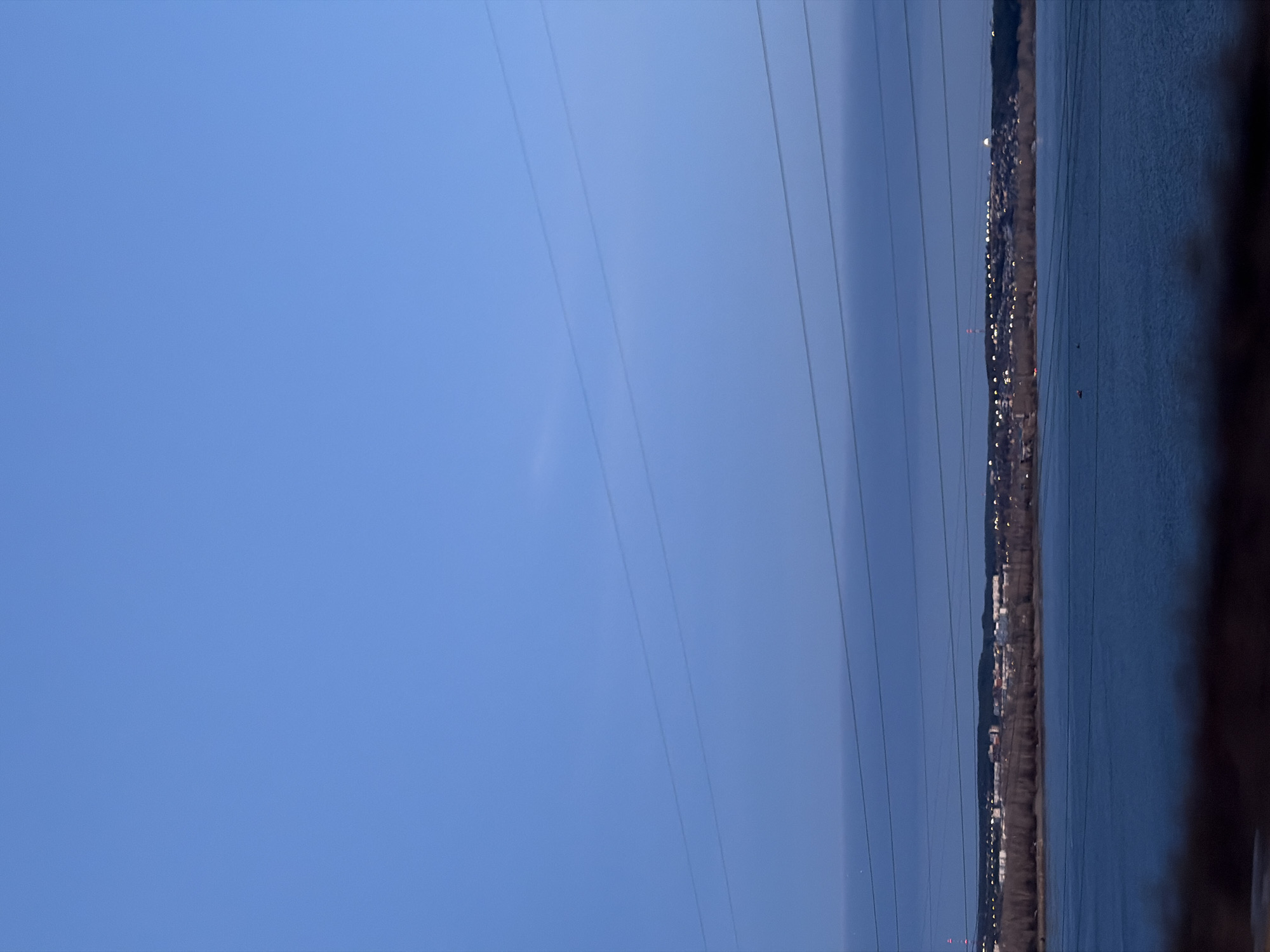 The image depicts a landscape with an expanse of water on the right side, adjacent to a stretch of land featuring buildings and structures illuminated by lights. Overhead, power lines traverse the sky, which transitions from a deep blue to a lighter hue. The scene appears to be taken near a body of water, possibly near the city of Chaikovsky, Russia, as indicated by the presence of urban infrastructure alongside the waterway. The image captures the interaction of the built environment with the natural landscape.