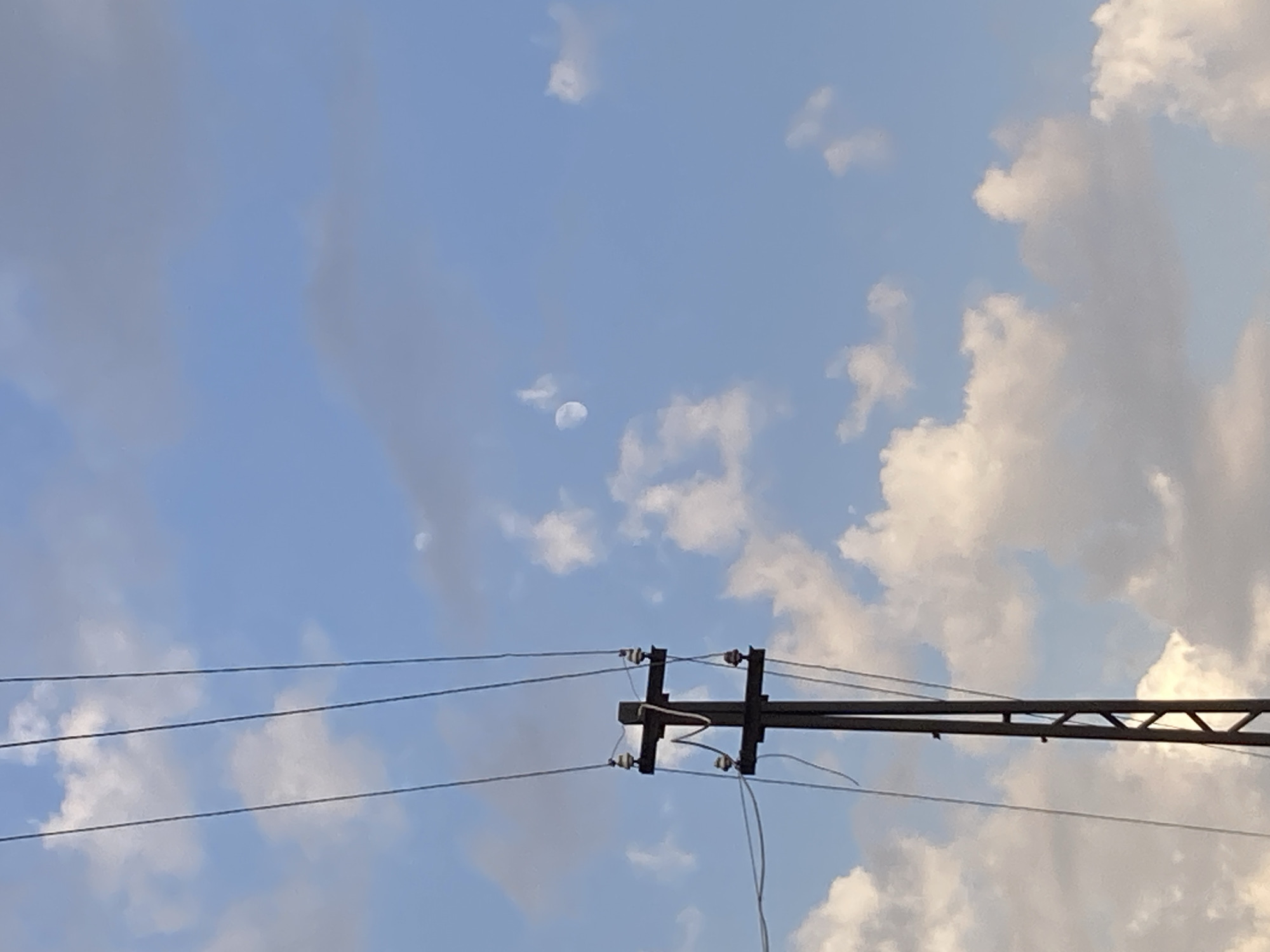 The image shows a utility pole with multiple wires against a blue sky with scattered white clouds. The pole is dark brown or black and has a crossbar structure with insulators. Several thin black wires are suspended from the insulators and stretch across the frame. In the upper right quadrant of the sky, a faint, partially visible moon is present. The overall scene is outdoors, suggesting an open area, possibly in the city of Avanos İlçe Merkezi, Turkey, given the context. There are no people or animals visible in the image.