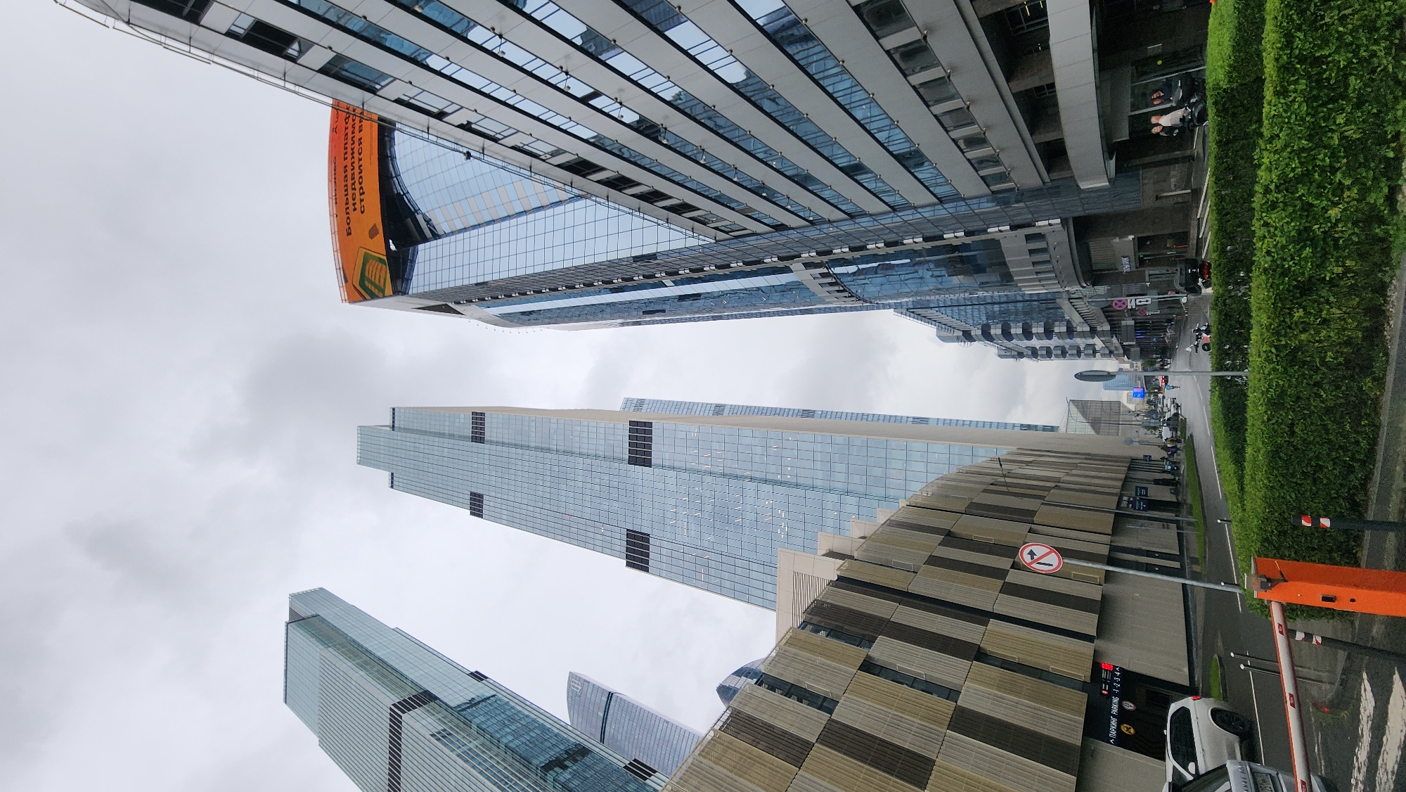 The image captures a view of modern skyscrapers in Moscow, Russia, on an overcast day. The camera is angled upwards, emphasizing the height and architectural design of the buildings, which are predominantly glass and metal structures.
In the foreground, a street with a road and a paved sidewalk is visible, lined with a dense green hedge. Various vehicles, including cars and motorcycles, are present on the road, suggesting an active urban environment. A white car is parked near a white and orange barrier, possibly at the entrance to a parking facility. A sign with a red circle and a diagonal line over an arrow indicates no entry.
The buildings themselves are a prominent feature. One large building on the left has a curved section with a large orange banner displaying text in Cyrillic: "Большая плата недвижимости строится" which translates to "Large payment of real estate is being built". Another tall, slender skyscraper dominates the mid-ground, its facade composed of reflective glass panels. Smaller buildings and structures are also visible in the distance, contributing to the dense urban landscape. The sky is a uniform gray, indicating cloudy weather. The overall impression is one of a bustling, contemporary city.