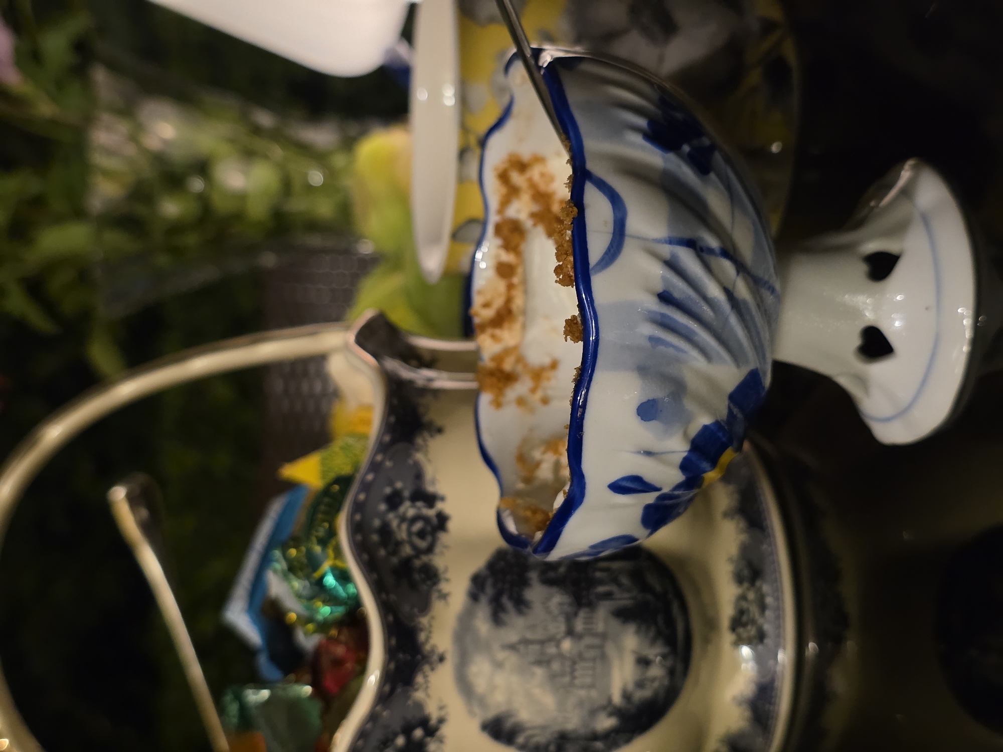 The image shows a dessert served in a decorative porcelain dish with blue and white patterns. The dish is on a table with a dark, reflective surface. In the background, there is a basket filled with colorful candies, a vase with greenery, and other dishes. The lighting is dim, creating a soft and intimate atmosphere. The image appears to have been taken indoors, possibly in a restaurant or home. There is no specific information about the location in Buzovna, Azerbaijan.