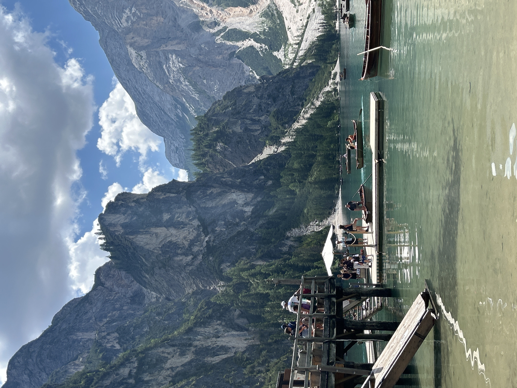 This media file depicts a picturesque mountain lake scene. The primary subjects are a group of people on a wooden pier extending into the clear, greenish water. Some individuals are seated, while others appear to be standing or about to board boats. Several small, wooden rowboats are visible in the water, some occupied by people with oars.
The setting is a dramatic mountain landscape, with steep, rocky slopes covered in dense green trees dominating the background. The sky is partly cloudy, with large white clouds against a bright blue expanse. Sunlight reflects off the water, creating shimmering patterns. The overall impression is one of a beautiful, serene day in a natural, mountainous environment, likely during daylight hours. The presence of people engaged in leisure activities suggests a vacation or recreational outing. There is a white umbrella on the pier, providing shade. Some of the people appear to be children or teenagers. A small sign with the numbers "015" is visible on the pier. The orientation of the image is vertical, implying it was taken with a phone or camera held in portrait mode.