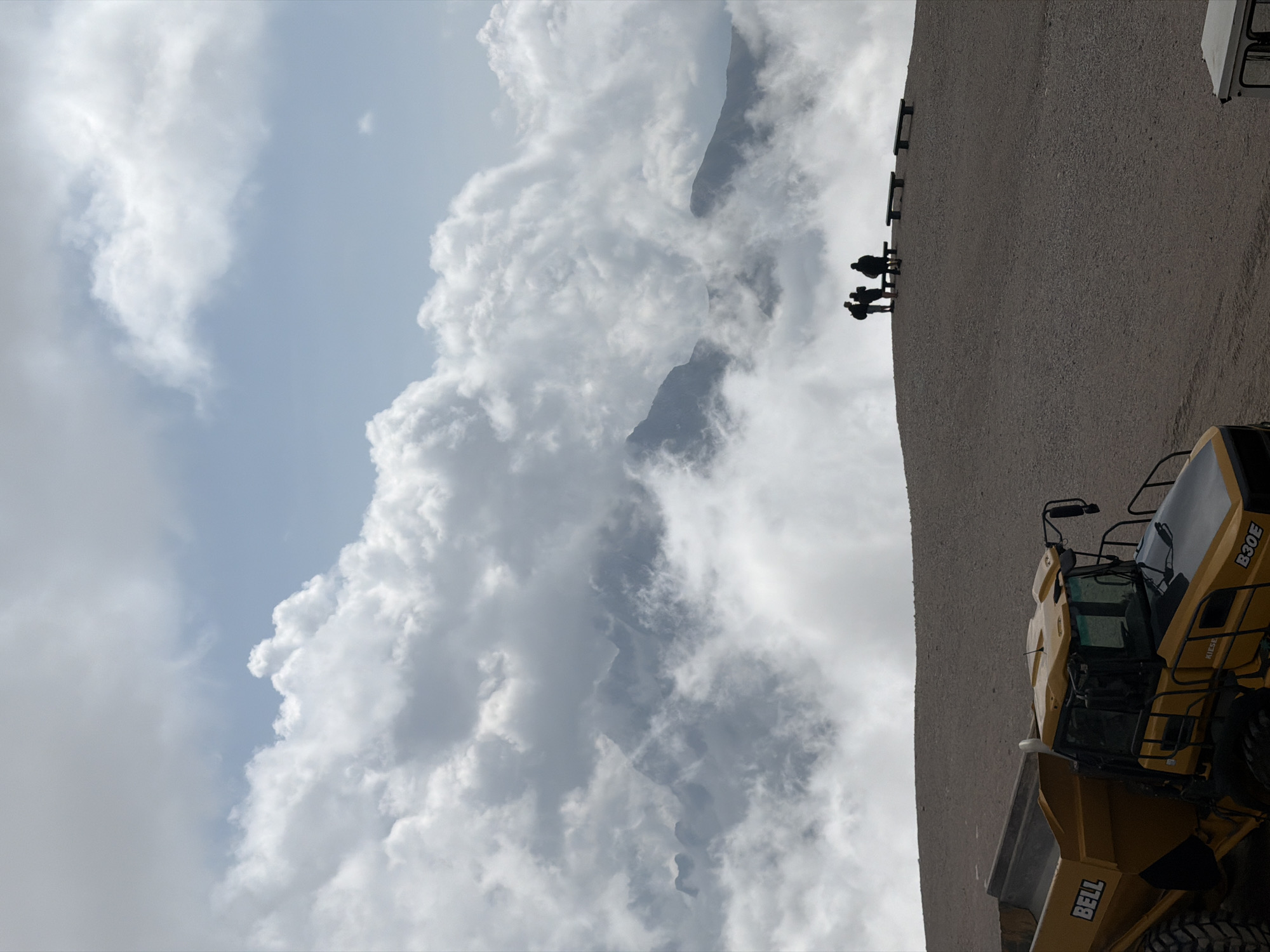 An image captures a dramatic mountain landscape shrouded in clouds, with a group of people silhouetted on a hilltop in the foreground. The scene is set in Garmisch-Partenkirchen, Germany, likely at a high altitude, given the cloud cover and rugged terrain.
In the foreground, a yellow Bell articulated dump truck is partially visible, suggesting construction or mining activity. Its large tires and robust frame indicate its industrial purpose. Behind the truck, the ground is covered in gravel, forming a steep incline.
Further up the incline, a group of four individuals stands near a set of three dark-colored benches. Their figures are small against the vast backdrop, emphasizing the scale of the mountains and clouds. They appear to be observing the panorama, their postures suggesting contemplation or awe. The clouds are dense and billowy, obscuring much of the mountain peaks, creating an ethereal and dramatic atmosphere. The sky above is a mix of blue and grey, with patches of sunlight breaking through the cloud cover.