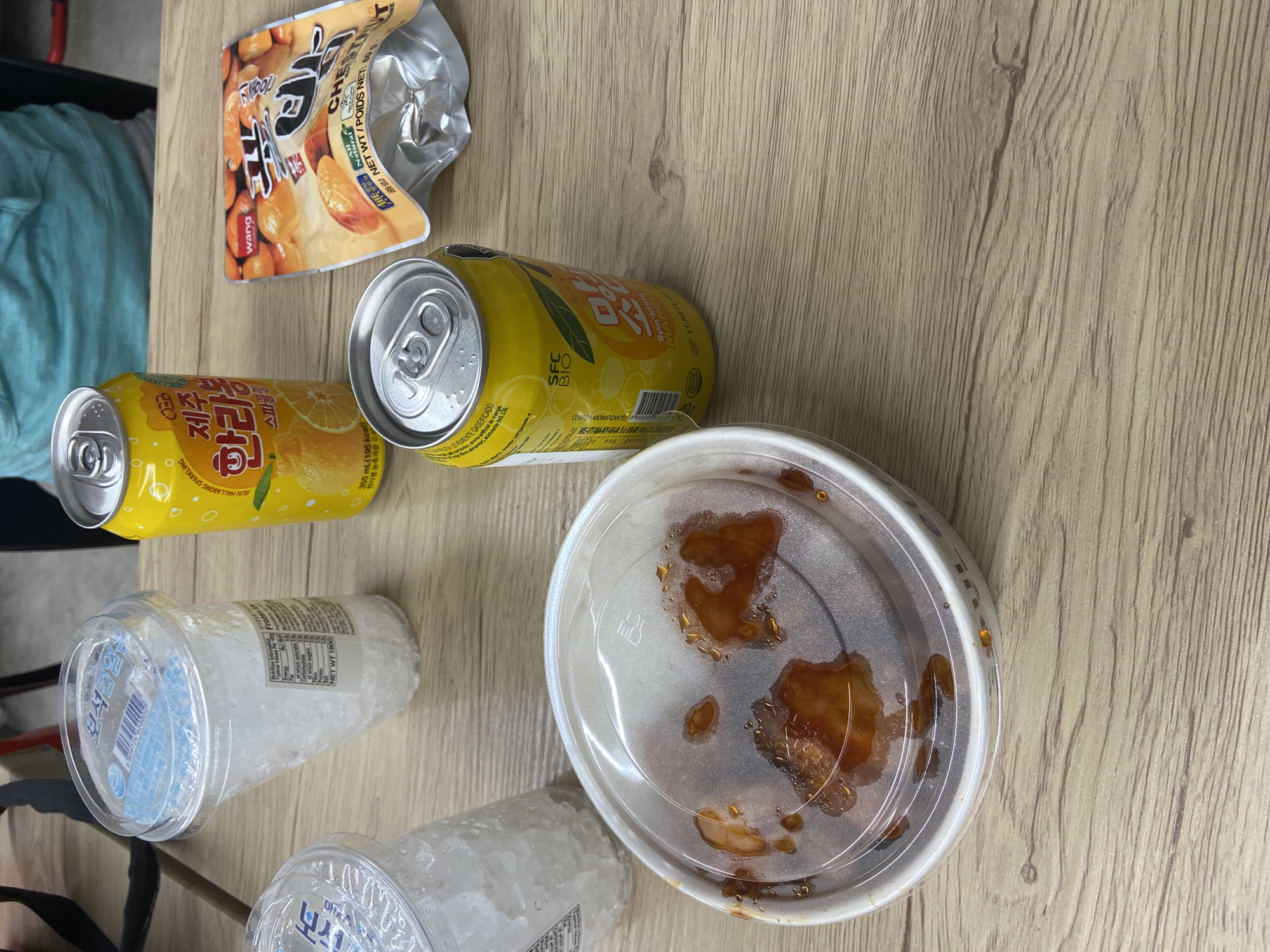 The image shows a table with various food and drink items scattered on it. There are two cans of Jeju Hallabong Sparkling drink, an opened bag of Wang chestnut snacks, a container with remnants of what appears to be fried chicken, and two cups containing a white substance, possibly ice or shaved ice. The table has a light wood grain finish. The overall impression is that of a casual, possibly finished, meal or snack setting. There is no information to suggest the location is in London.