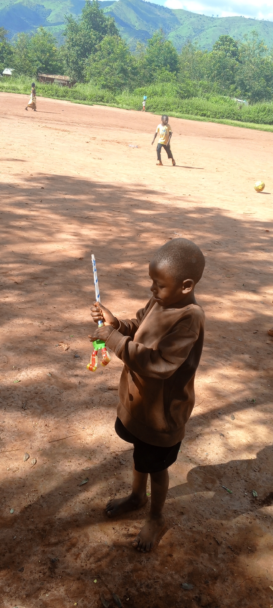 The image captures an outdoor scene in Ngalula, Democratic Republic of the Congo, on a sunny day. In the immediate foreground, a young, dark-skinned child, barefoot, wears a brown long-sleeved shirt and dark shorts. The child is holding a blue and white striped stick-like object, which features a green and yellow attachment at its lower end, and appears focused on this item.
The midground consists of an expansive, reddish-brown dirt field, characteristic of an unpaved open space. Several other children are present in this area: one child in light-colored attire walks from the right to the left on the far left of the frame; another child, wearing a yellow t-shirt and dark pants, is walking towards the right; and a fourth child in light blue clothing is faintly visible further back. A yellow football rests on the dirt ground on the right side of the midground. Shadows, indicative of overhead sunlight, are cast across the ground.
The background is dominated by a line of dense green trees and vegetation, situated beyond the dirt field. Behind these trees, a range of large, verdant mountains rises under a clear to partly cloudy blue sky. A small, partially obscured structure with a visible rusty roof is discernible behind the trees on the left side of the background.