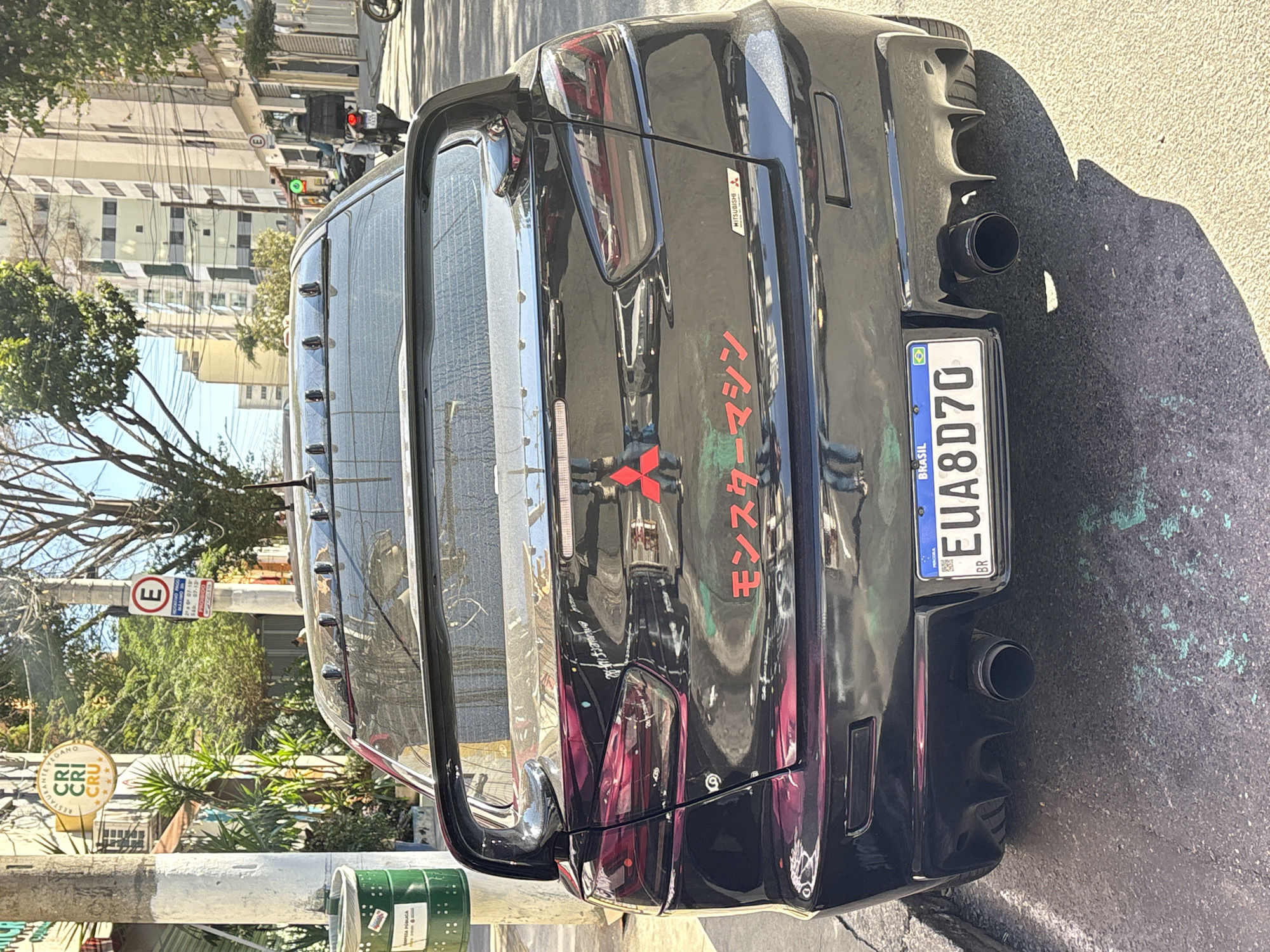 The image shows the rear of a black Mitsubishi Lancer Evolution parked on a street in São Paulo, Brazil. The car has a large rear spoiler and a custom decal on the trunk that reads "モンスターマシン" (Monster Machine) in Japanese characters, along with the Mitsubishi logo. The license plate is visible and reads "EUA8D70". A green trash can with a sign is on the left side of the image, and a traffic sign indicating parking restrictions is visible on a pole. Buildings and trees line the street, and a motorcycle is seen in the background. The scene appears to be a sunny day.