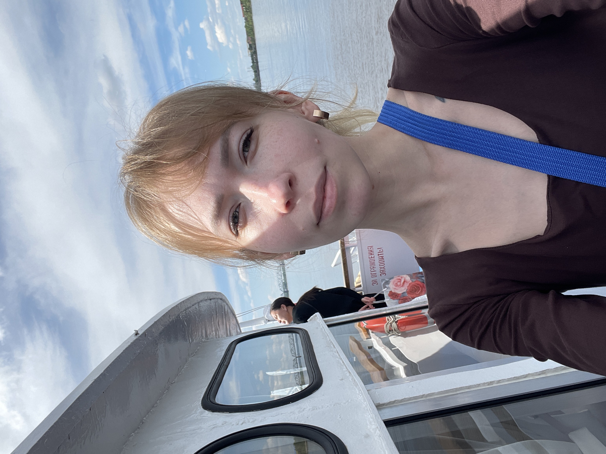 The image is a selfie of a young woman taken on a boat or ship, capturing her face and the scenic background. The woman, with light blonde hair, fair skin, and freckles, looks directly at the camera with a neutral expression and a slight smile. She wears a dark brown long-sleeved top and a prominent blue strap across her chest and shoulder. A small gold-colored rectangular earring is visible on her right ear, and a faint dark mark, possibly a tattoo, is on her left shoulder.
The setting appears to be a river cruise on a sunny, partly cloudy day. Behind the woman, the calm blue water of a wide river stretches into the distance, with a distant green shoreline and some buildings visible. The bright blue sky above is dotted with white clouds.
The boat itself features white and light grey painted structures, including a railing and windows. Further back on the boat, another individual with dark hair and wearing a dark top is visible, leaning or seated near a red lifebuoy wrapped with white rope. A white sign with illustrations of pink roses and the Russian text "ЗА ОГРАНИЧЕНИЯ ЗАХОДИТЬ!" (translating to "ENTER FOR RESTRICTIONS!") is also partially visible on the boat. The scene suggests a relaxed outdoor activity, likely on the Kama River in Perm, Russia.