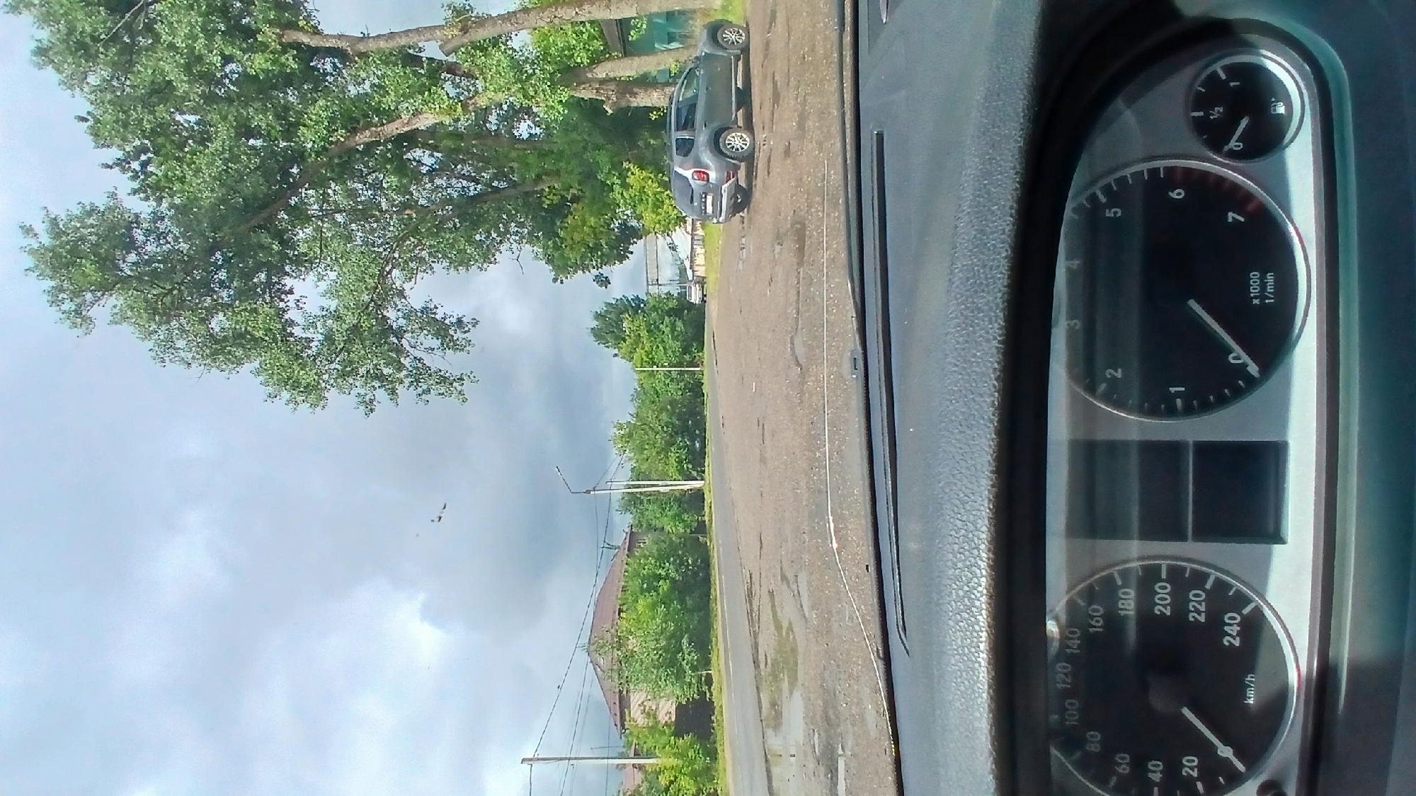 The image, taken from inside a vehicle in Mozhaisk, Russia, shows a view of a paved area with puddles, likely a parking lot or road, under a cloudy sky. Large trees with green foliage line the right side of the frame. A grey Dacia Duster SUV is parked on the right, facing away from the camera. Further in the distance, another vehicle, possibly a white van, is visible near a structure with a metal frame. The foreground displays the car's dashboard with a speedometer indicating a speed of approximately 20 km/h, and a tachometer showing just under 1000 RPM. Utility poles and wires are present on the left side, along with houses and dense green trees in the background. The overall scene suggests an overcast day in a semi-urban or suburban area.