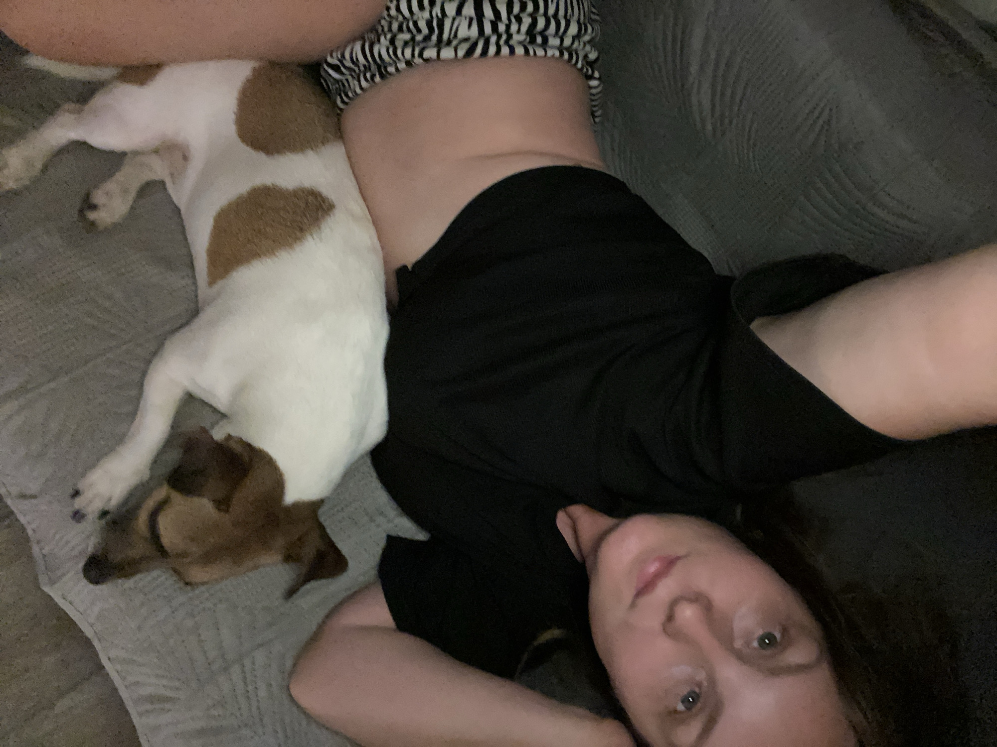 The image depicts a low-angle, close-up shot of a woman and a small dog lying on a textured gray sofa. The woman, positioned on the right side of the frame, is lying on her back with her head propped on her arm, looking towards the camera. She has brown hair, light skin, and is wearing a black t-shirt and patterned shorts with black and white stripes. The dog, a white and brown Jack Russell Terrier, is lying on its side on the woman's lower torso and legs. The dog has its eyes closed and appears to be sleeping. The background is dominated by the gray sofa, with some indication of a darker surface behind the woman's head. The scene appears to be indoors, likely in a living room setting. There is no visual information in the image that allows for the determination of its location within the city of Noviy, Russia.
