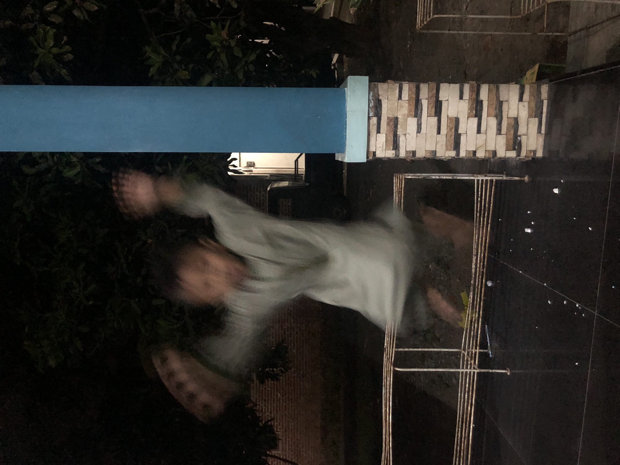 A child, appearing blurred due to motion, is captured mid-jump outdoors at night, likely in Tenggilis Rejo, Indonesia. The child is wearing a light-colored, loose-fitting garment, possibly a traditional outfit, with arms outstretched. They are positioned in front of a dark background of foliage and trees, with a brick wall visible to the left and a blue cylindrical pillar with a decorative stone base to the right. In the mid-ground, a light-colored metal railing with multiple horizontal bars runs across the frame. The foreground consists of a dark, reflective tiled surface. A portion of a vehicle is partially visible behind the child and the railing. The lighting suggests artificial light sources are present, creating a dimly lit, atmospheric scene.