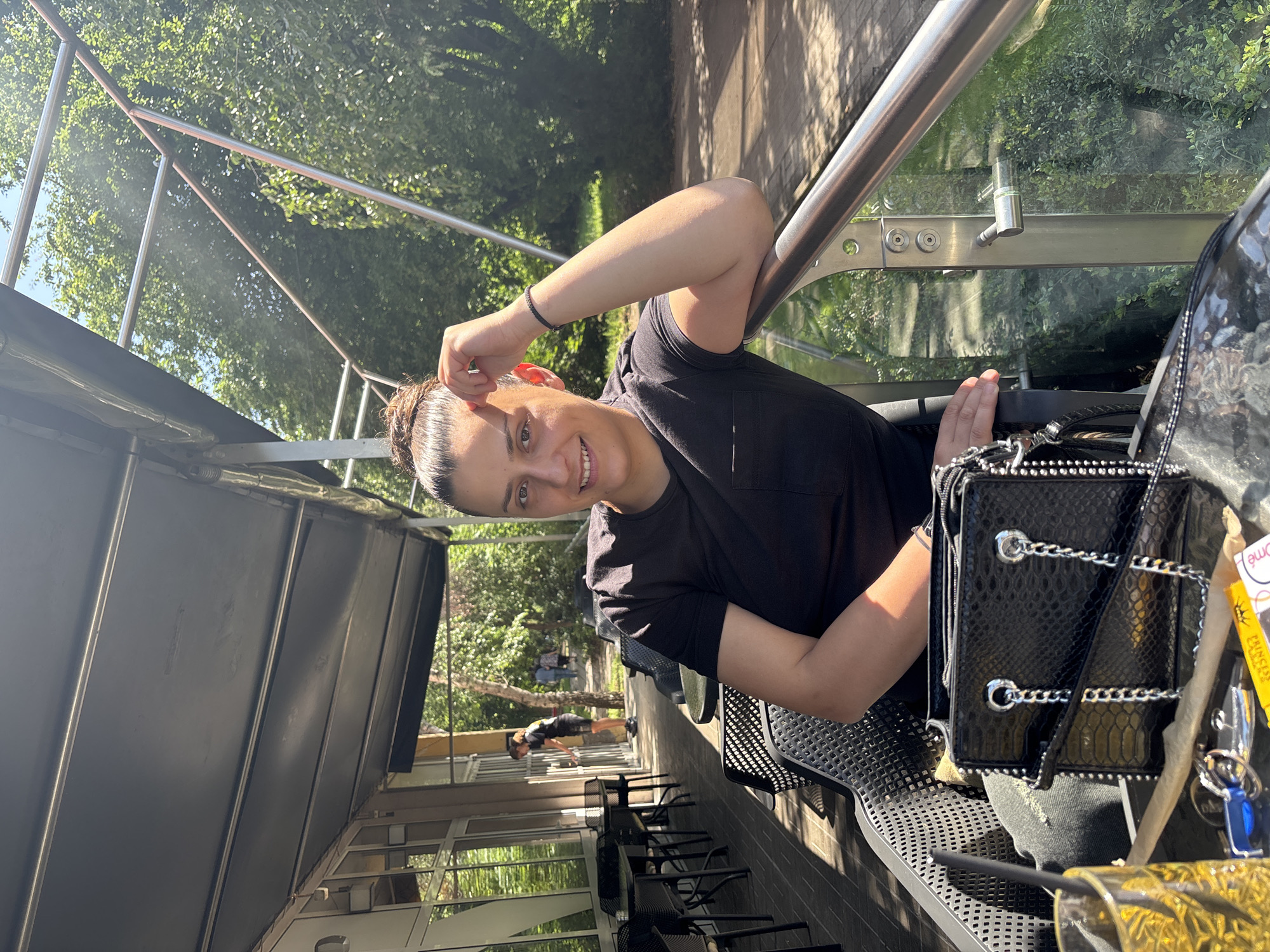 A bright, sunlit, outdoor scene shows a person (likely a woman) smiling at the camera, seated at what appears to be a cafe or restaurant in Sofia, Bulgaria. The subject has dark hair pulled back, wears a black t-shirt, and has a black beaded bracelet on their left wrist. Their right arm rests on a metal railing.
On the table beside them is a black bag with silver chain straps and a snakeskin-like texture. Next to the bag are keys with a bright blue fob and a yellow tag. A clear plastic cup with a black straw and a yellow-patterned label is also visible on the table.
The setting is an outdoor seating area, possibly under an awning, indicated by the shaded foreground and bright background. Black metal chairs and tables are arranged along a brick-paved area. In the mid-ground, a few other blurry figures can be seen walking or standing near tables. Beyond the seating area, lush green trees and foliage fill the background, suggesting a park or tree-lined street. A concrete wall or path runs through the greenery.
The time of day is sunny daytime, with bright natural light and visible shadows. The subject appears happy and relaxed, directly engaging with the viewer. Partially visible text on the yellow tag next to the keys includes "PRINCESS" and "eome". The overall atmosphere is casual and pleasant.