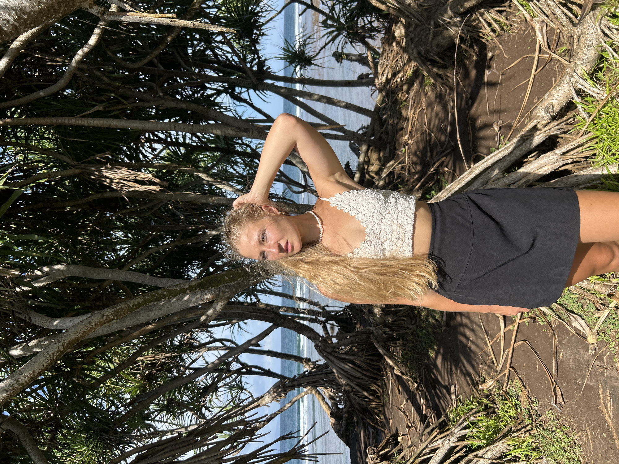 A young woman with long blonde hair poses in a wooded area with the ocean visible in the background, which is identified as Kuta Selatan, Indonesia. She wears a white lace top, a black skirt, and a pearl necklace. Her left hand is raised, touching her hair. The environment is characterized by the presence of trees with visible roots and branches. The ground is dirt with some vegetation. In the background, the ocean meets the horizon under a blue sky.