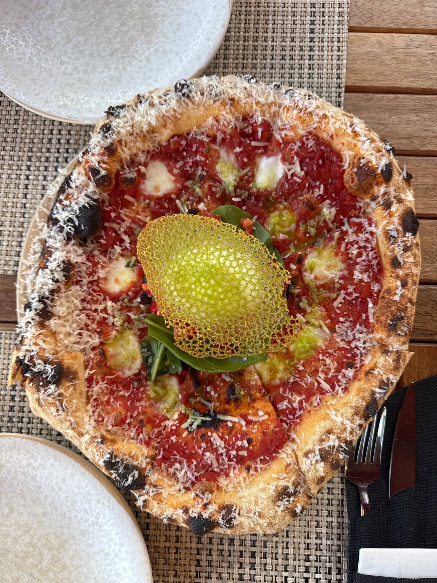 An overhead shot captures a round pizza with a thick, charred crust, generously topped with tomato sauce, melted white cheese, and grated Parmesan. A distinctive bright yellow, lacy garnish sits atop a small cluster of green leaves in the center of the pizza. The pizza rests on a woven placemat on a wooden table. To the right of the pizza are a fork and knife placed on a black napkin, and a small white napkin. Partially visible in the upper left and lower left corners are white plates with textured rims. The setting appears to be an outdoor dining area, likely in Konyaaltı, Turkey, given the proximity to the Mediterranean coast and the commonality of such establishments there. The scene suggests a meal is about to be enjoyed.