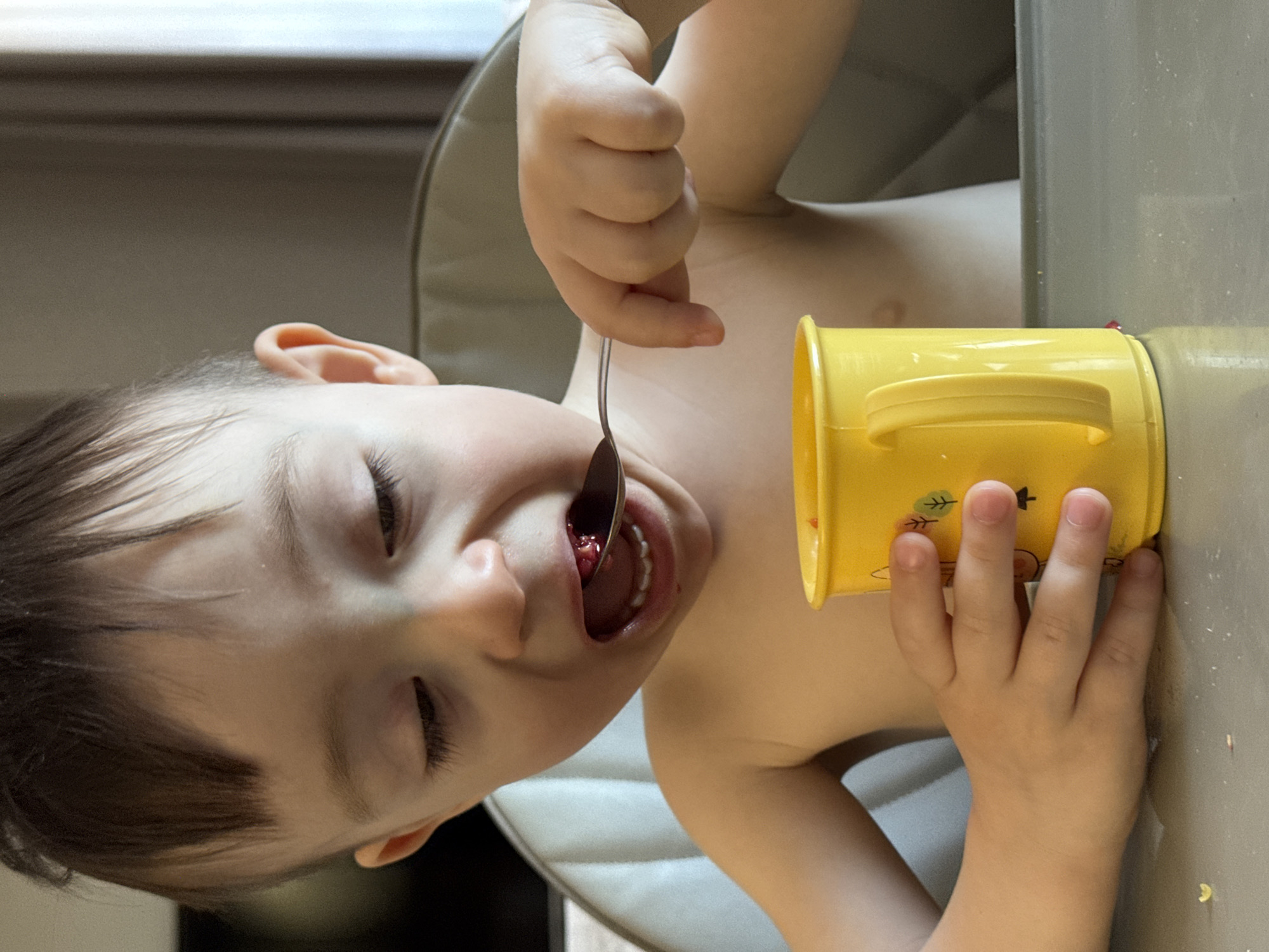 A young child is shown from a close, angled perspective. The child is holding a yellow cup in their right hand and a spoon in their left, placing food into their mouth. The location and city are not discernible from the image.