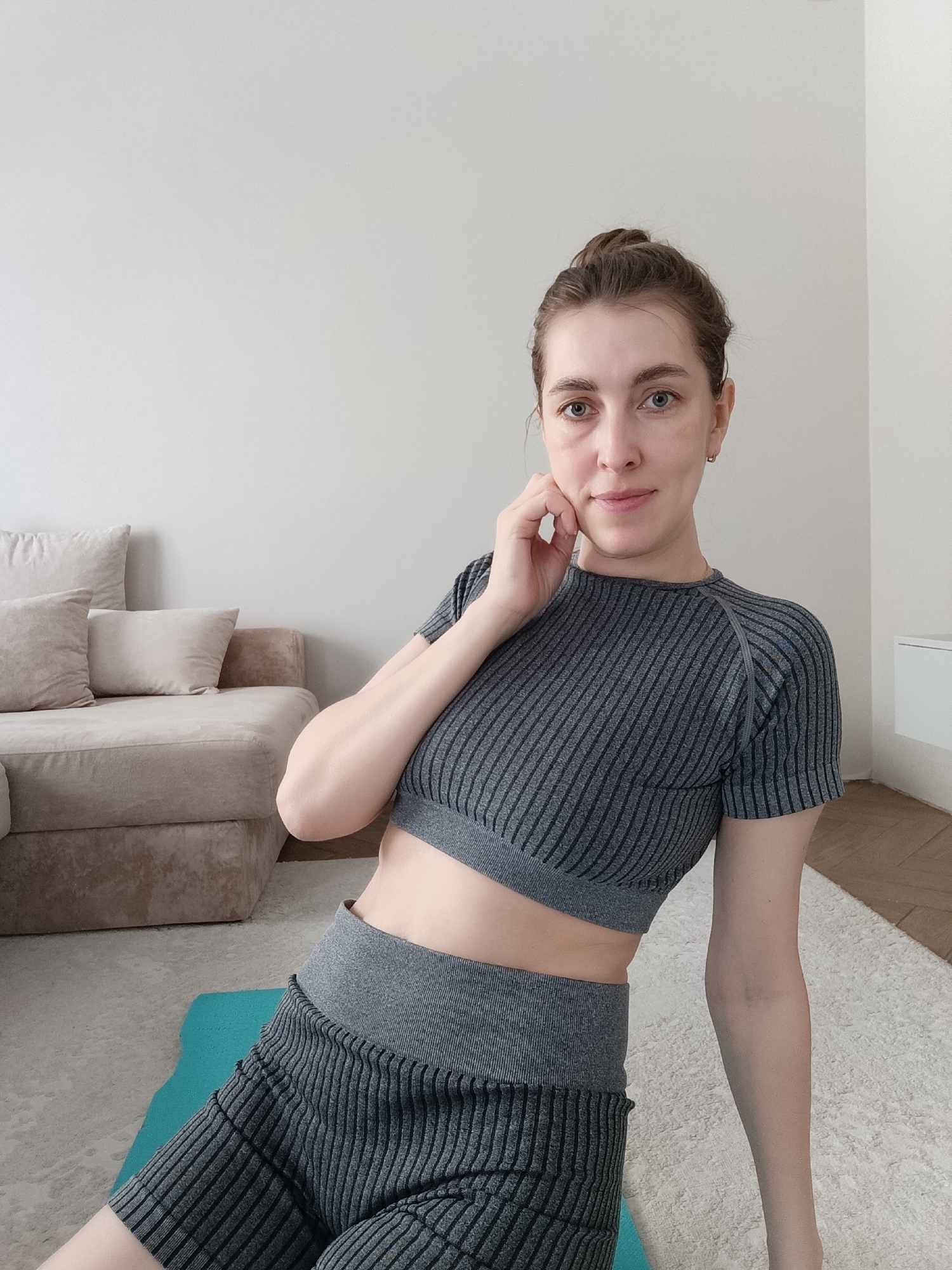 A woman is shown in a medium indoor shot, seated on a blue yoga mat that is placed on a white rug. She is wearing a grey, striped exercise outfit consisting of a crop top and shorts. She is partially leaning on her left hand, which is placed near her face. Her hair is pulled back into a bun. Behind her is a light-beige couch with pillows and a white wall. A corner of a wooden floor can be seen on the right side of the rug. The location is likely Chaikovsky, Russia.