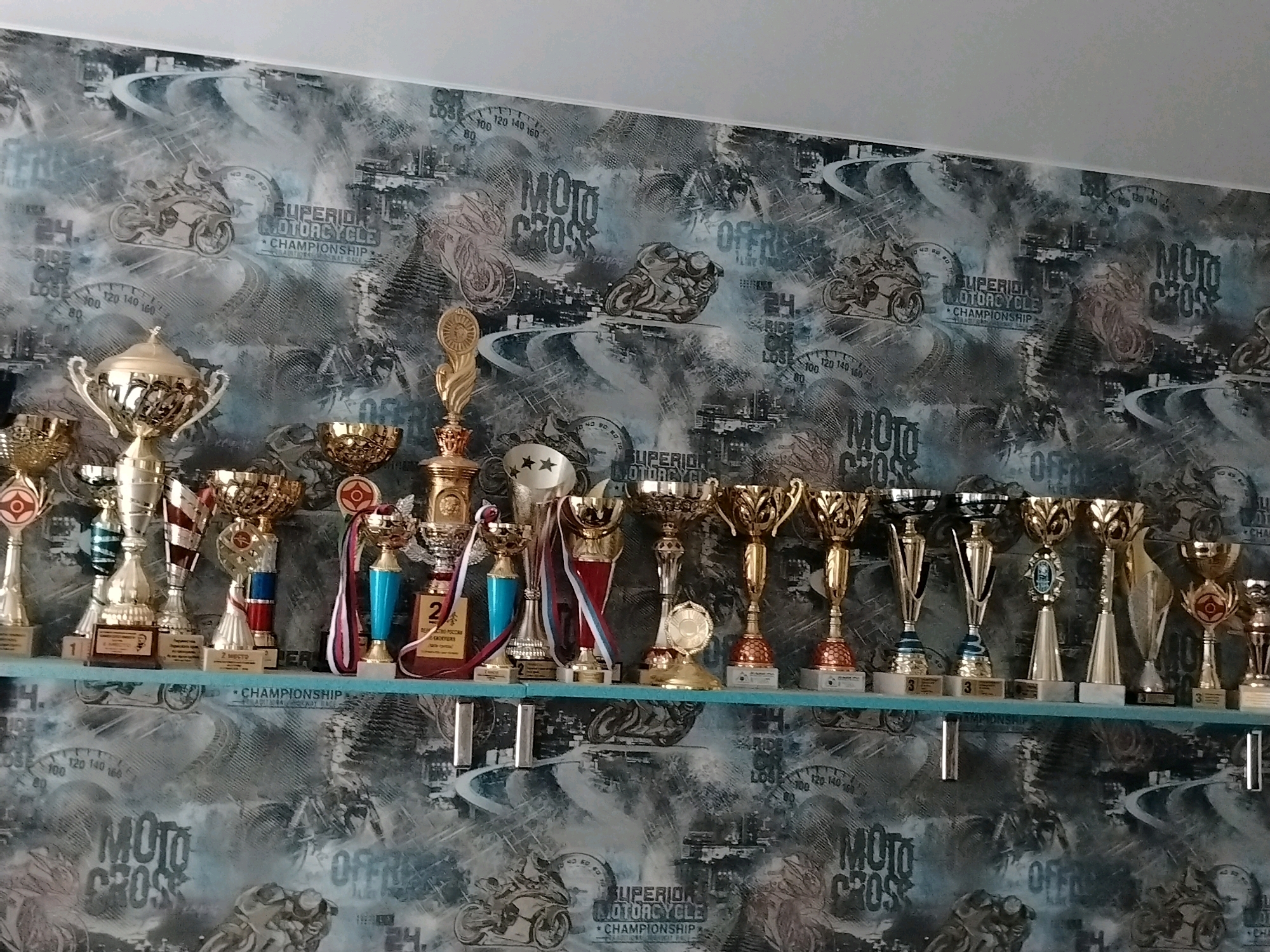 The image displays a collection of trophies arranged on a shelf against a motorcycle-themed wallpaper. The trophies are of various sizes and designs, predominantly gold and silver in color, some adorned with ribbons. The wallpaper features a collage of motorcycles, speedometers, and text related to motocross and championships. The scene appears to be an indoor setting, possibly a room or a display area. The photograph is taken from a slightly elevated angle, looking down at the trophies. There are no people visible in the image. The trophies are the central focus of the image. The environment suggests a space dedicated to celebrating achievements in motorsports.