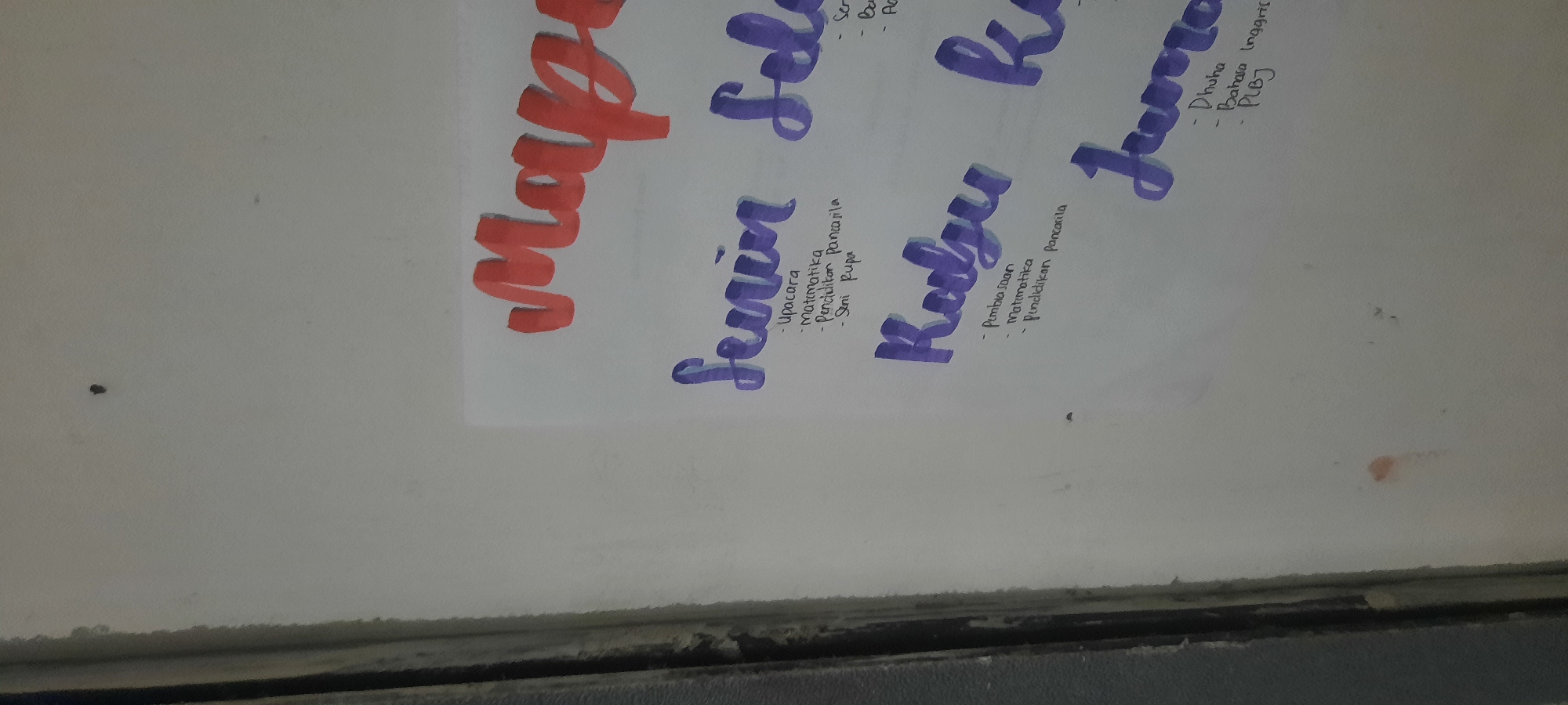 The image shows a white wall with handwriting on it. The handwriting is in a colorful script, with the days of the week written in large, bold letters. Underneath each day of the week, there is a list of subjects or activities. The subjects listed appear to be school-related. There are also some small stains on the wall. The image is taken from a slightly upward angle, and the lighting is somewhat dim. The scene does not provide enough information to determine the specific location in North Jakarta, Indonesia.