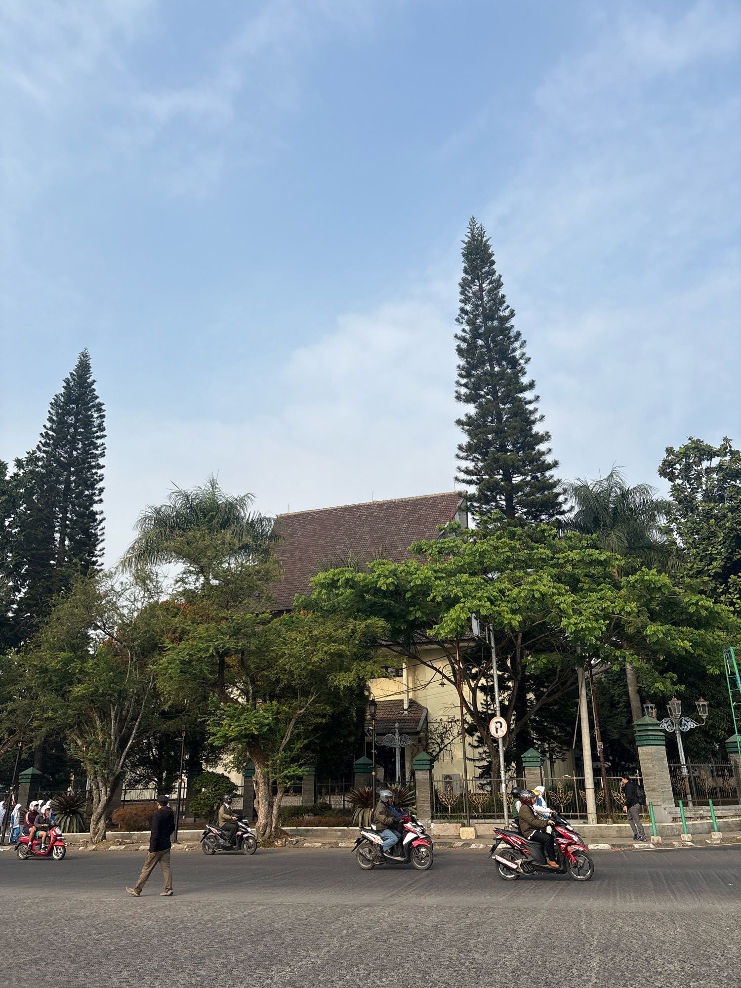 The image depicts a street scene in Pamekasan, Indonesia, featuring a large building with a brown tiled roof partially obscured by lush green trees. Two tall, slender pine trees stand out against a pale blue sky with scattered clouds. Several individuals are present, with some riding motorbikes on the paved road. One person is walking across the street, and others are standing near the side of the road. A traffic sign with a "P" symbol is visible on a pole. The environment appears to be a city street with landscaping and architectural elements.