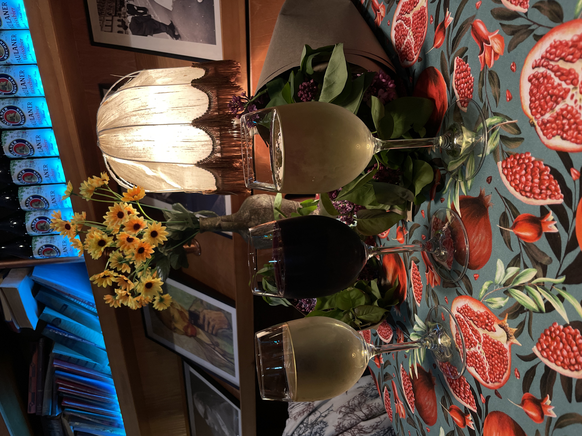 The image shows a scene at what is likely a wine bar or restaurant, potentially in Tbilisi, Georgia. The tablecloth features a vivid pomegranate design. Three wine glasses, filled with a white, red, and white liquid respectively, are arranged on the tablecloth. A bouquet of flowers and greenery, including sunflowers and what appear to be purple lilac, lies between the glasses.
Behind the glasses, a vintage lamp with a scalloped shade illuminates the scene. A bookshelf is also visible, stacked with books that are lit with blue light. Bottles of Paulaner Weissbier are arranged atop the bookshelf. Framed pictures hang on the wall.
