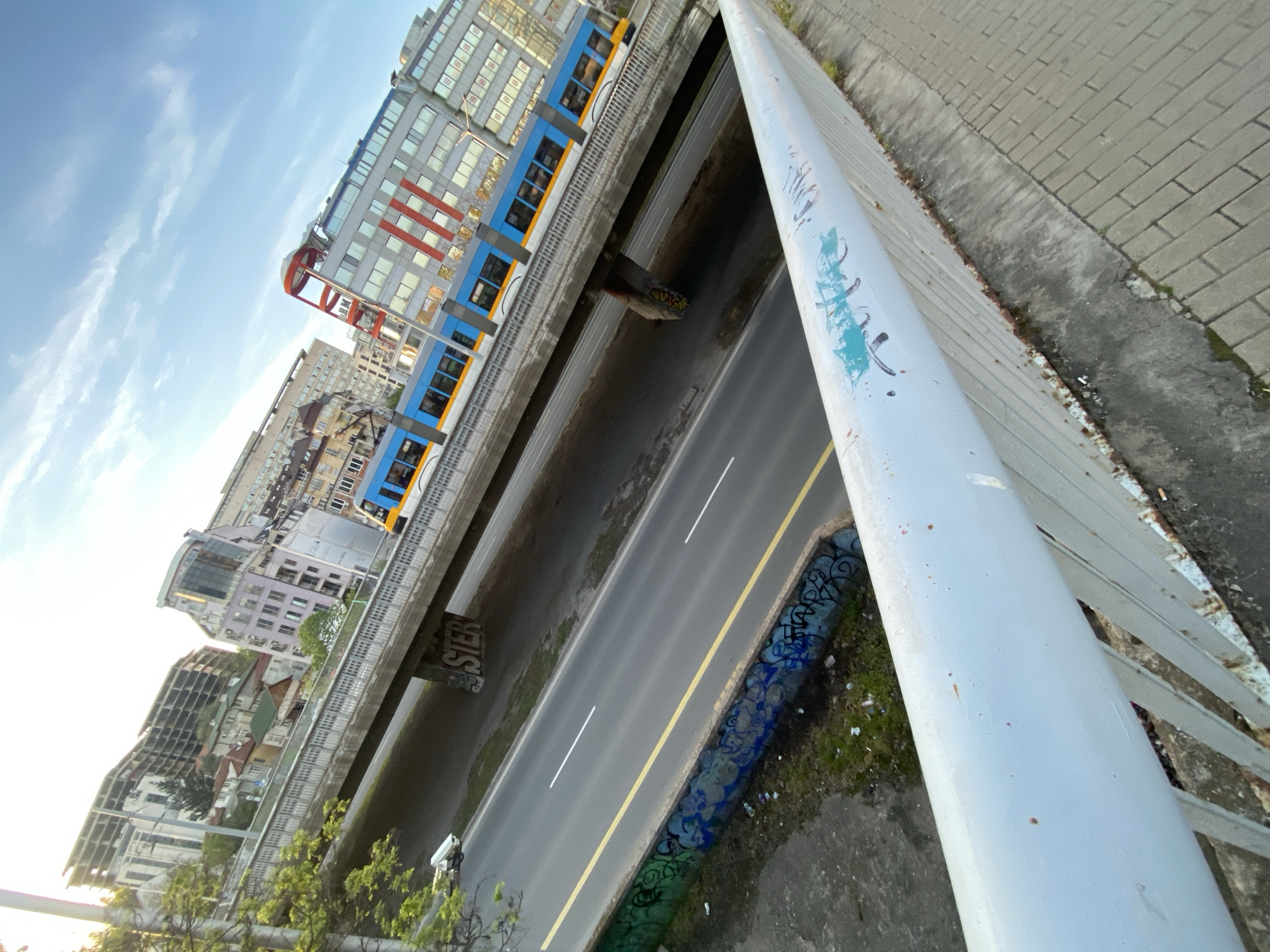 The image presents a diagonally tilted, elevated view of an urban landscape in Sofia, Bulgaria, looking down onto a street and an elevated train line. In the immediate foreground, a white metal railing, marked with blue and black graffiti, cuts across the right side of the frame.
Below the vantage point, a multi-lane asphalt road stretches horizontally, featuring a solid yellow median line and broken white lane markers. A small, white vehicle, likely a scooter or motorcycle, with a rider, is visible on the road. Adjacent to the road, a concrete barrier is covered extensively with blue and black graffiti.
Above the road, an elevated concrete track carries a modern blue and yellow train or metro car, which appears to be in motion. One of the concrete support columns beneath the elevated track has graffiti that clearly spells "SIER".
The background reveals a dense urban environment with a mix of architectural styles, from contemporary glass and concrete buildings with red accents to older, more traditional structures. The sky above is a bright, clear blue with a few scattered, wispy white clouds, indicating daytime. The bright lighting and reflections suggest it is likely late morning or afternoon.